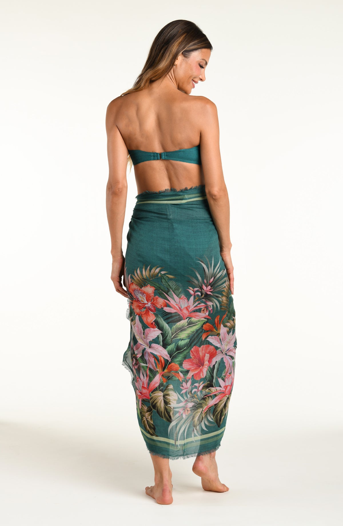 A model wearing a dark teal green tropical print bandeau bikini top and matching canga wrap skirt with a side slit, shown from the waist down against a white background.