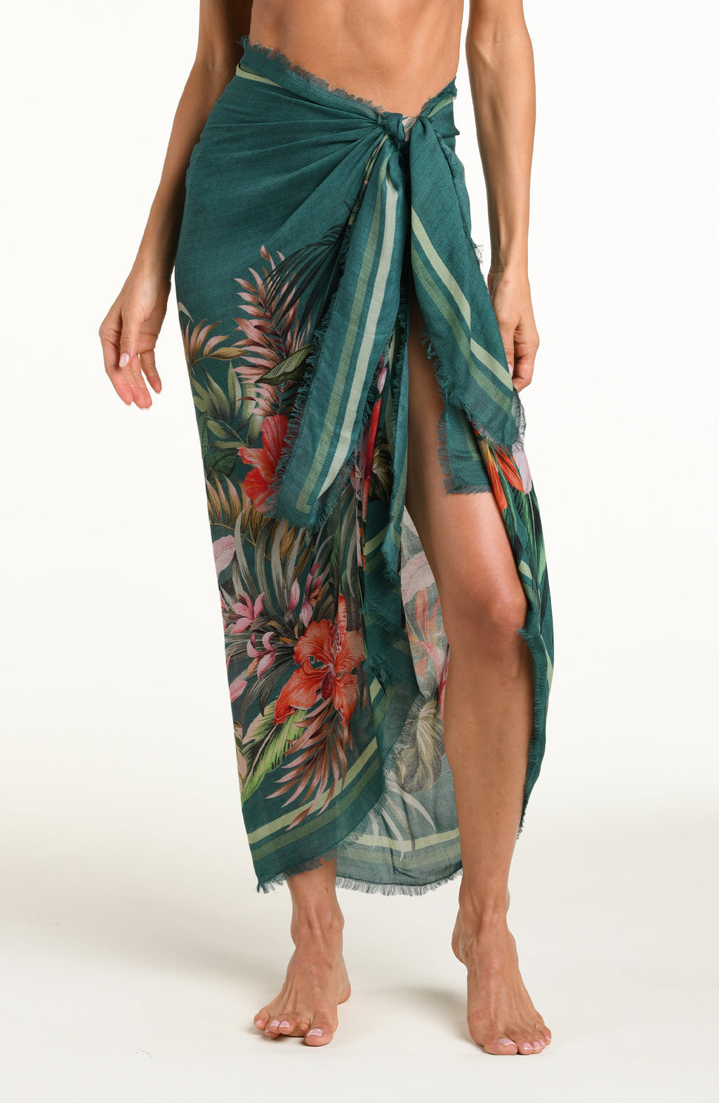A model wearing a dark teal green tropical printed canga wrap skirt with a side slit, shown from the waist down against a white background.