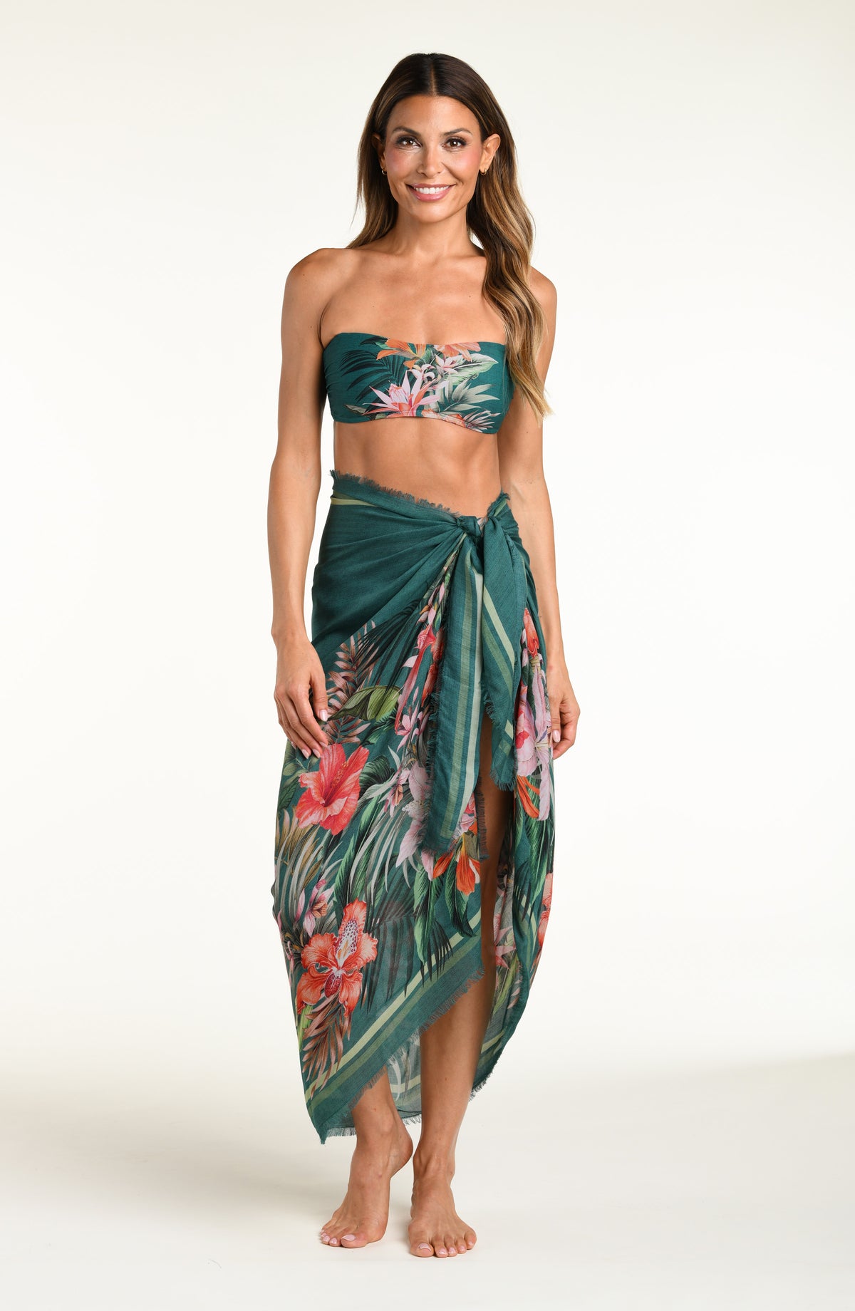 A model wearing a dark teal green tropical print bandeau bikini top and matching canga wrap skirt with a side slit, shown from the waist down against a white background.