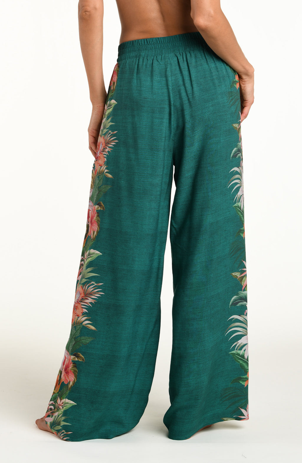 A model wearing wide-leg pants in a deep teal green color, featuring a large, vibrant tropical floral print with pink, white, and orange flowers and green leaves running down the side against a white background. 
