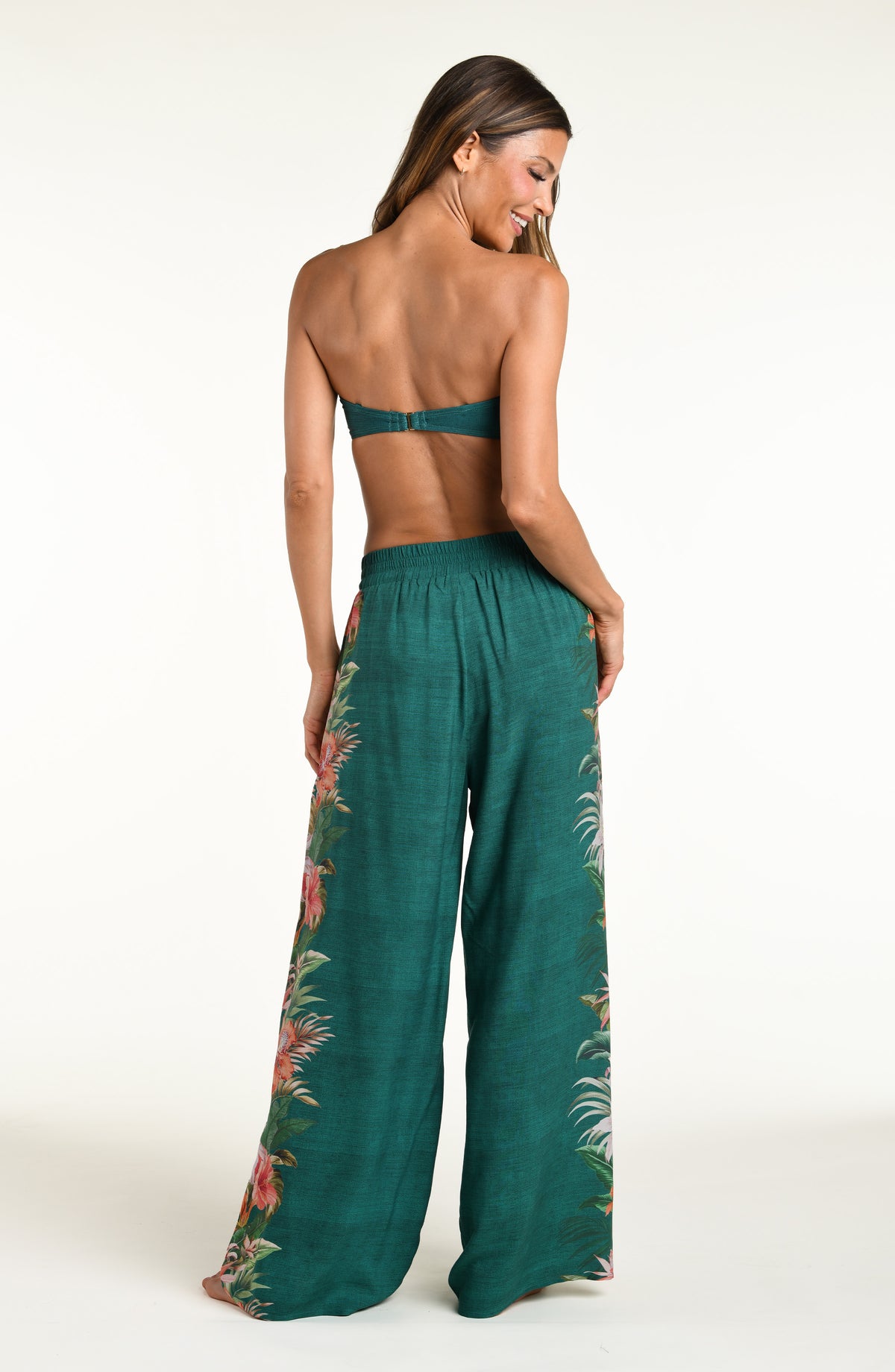 A model wearing a bandeau bikini top and wide-leg pants in a deep teal green color, featuring a large, vibrant tropical floral print with pink, white, and orange flowers and green leaves running down the side against a white background. 