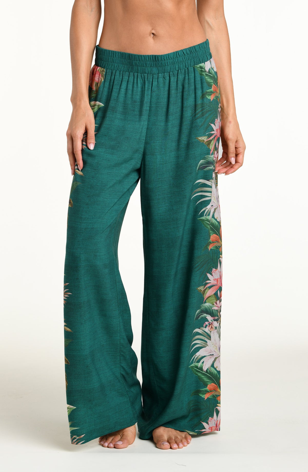 A model wearing wide-leg pants in a deep teal green color, featuring a large, vibrant tropical floral print with pink, white, and orange flowers and green leaves running down the side against a white background. 