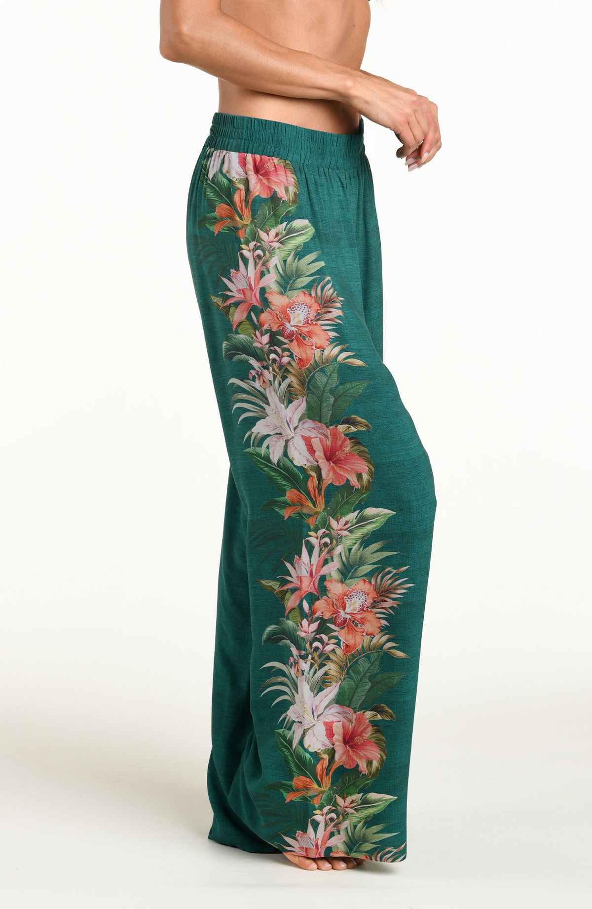 A model wearing wide-leg pants in a deep teal green color, featuring a large, vibrant tropical floral print with pink, white, and orange flowers and green leaves running down the side against a white background. 