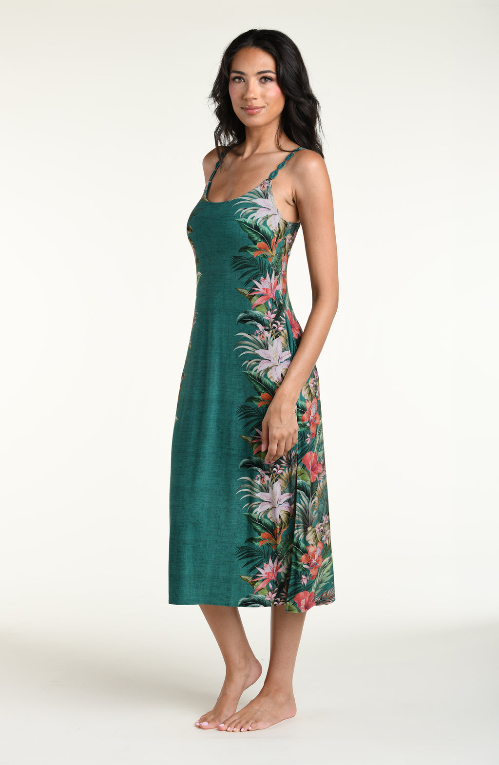 A woman modeling a sleeveless midi dress with thin straps and a tropical floral print in green, pink, orange, and white, photographed against a plain light background.