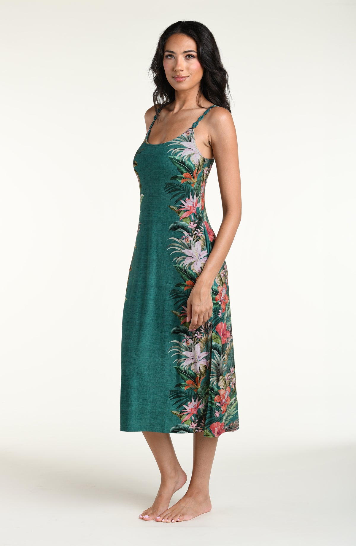 A woman modeling a sleeveless midi dress with thin straps and a tropical floral print in green, pink, orange, and white, photographed against a plain light background.