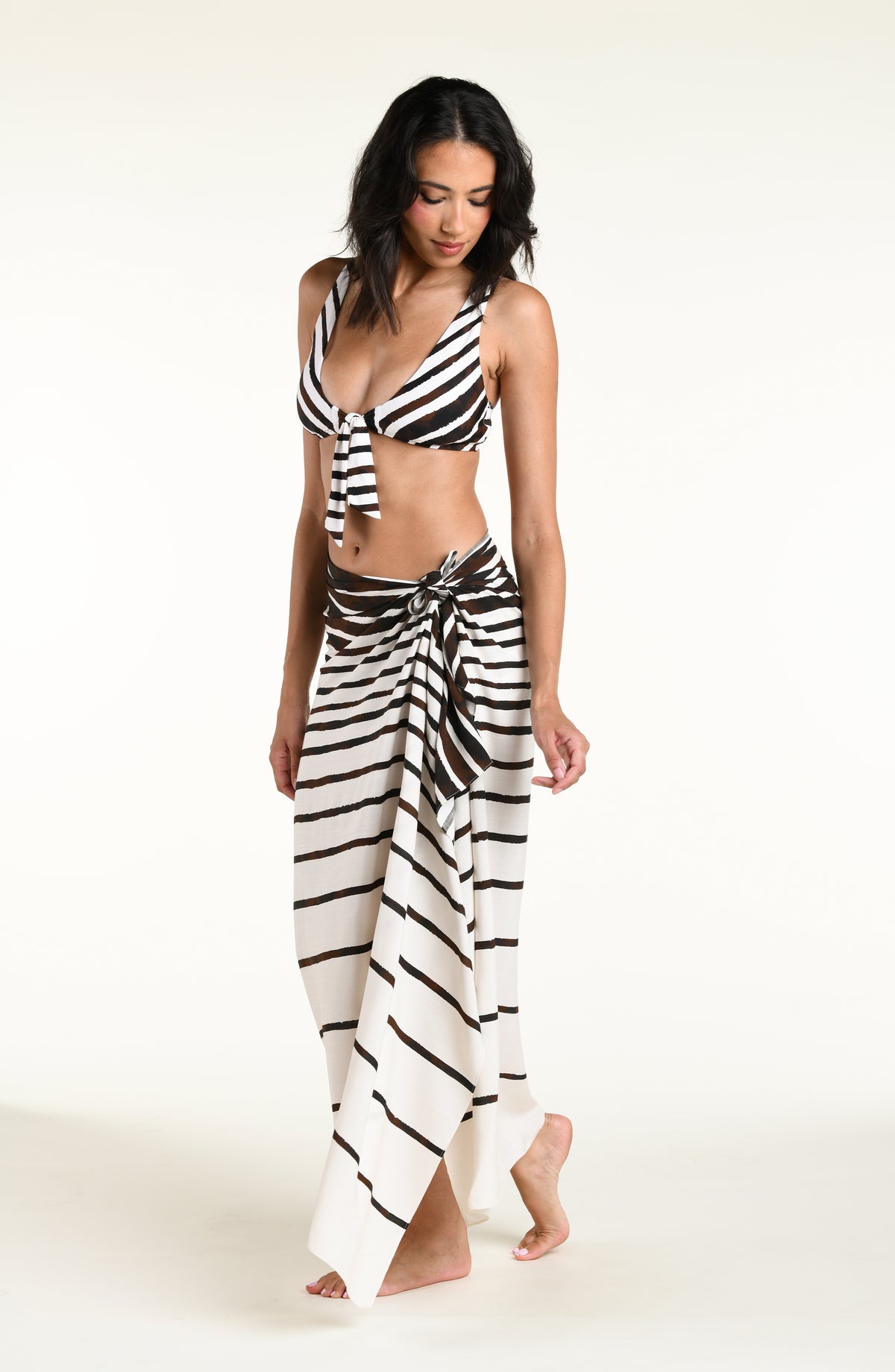A woman modeling a brown and white striped bikini top with a front tie and matching striped sarong wrap skirt, set against a plain white studio background.