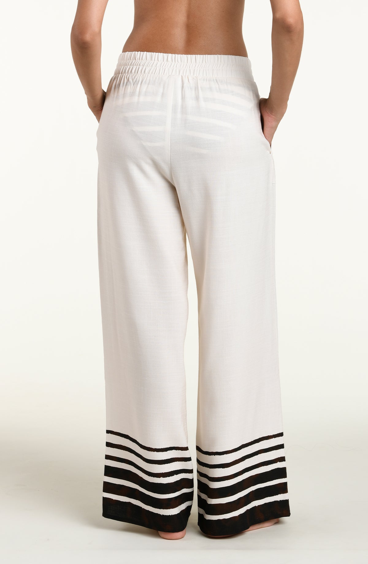 A woman wearing loose white wide-leg pants with brown horizontal stripes near the hem and a side pocket, shown against a plain light beige studio background.