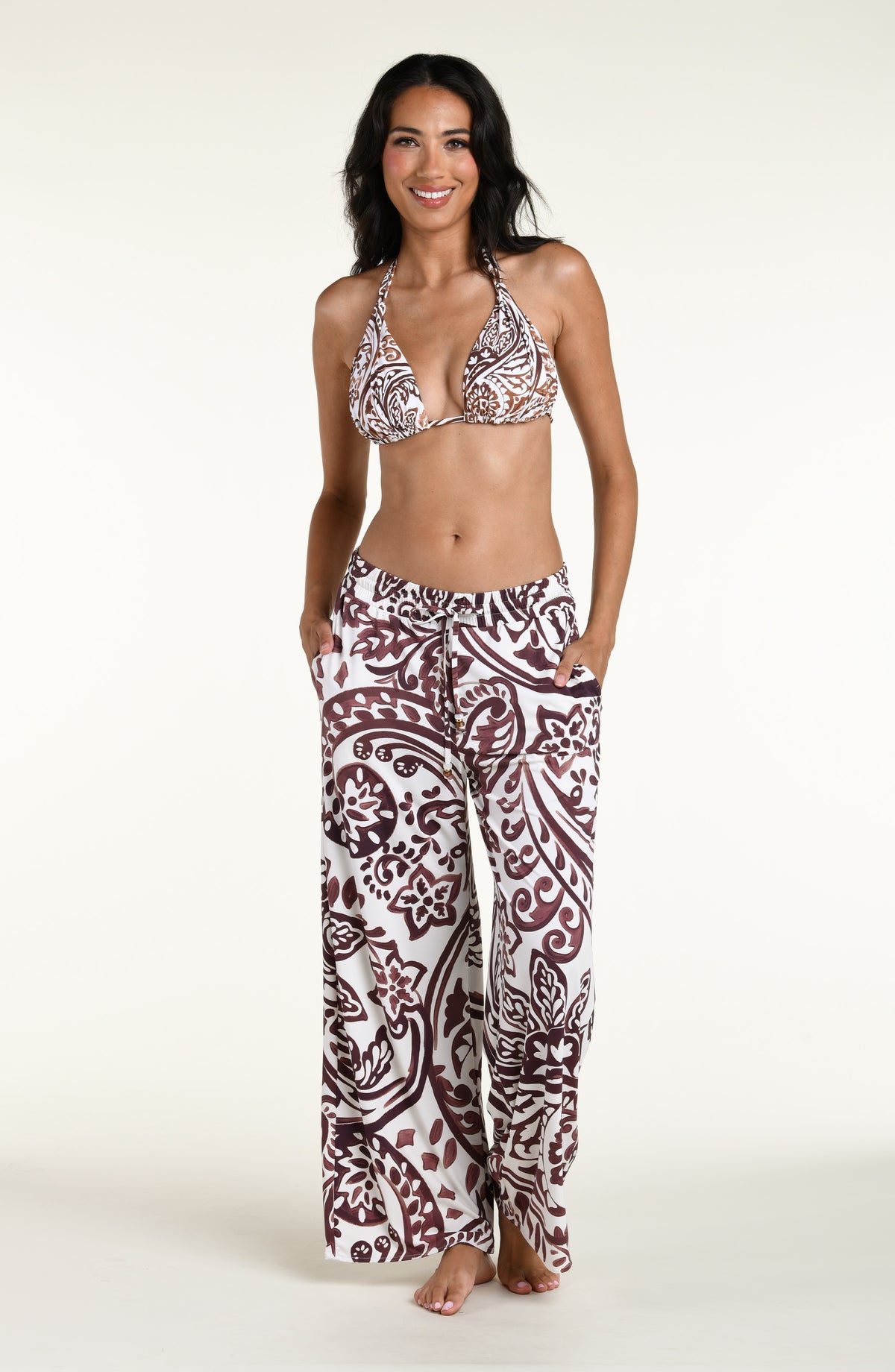 A woman modeling a bikini top and matching beach pants in a brown and white paisley pattern, standing against a plain off-white studio background.