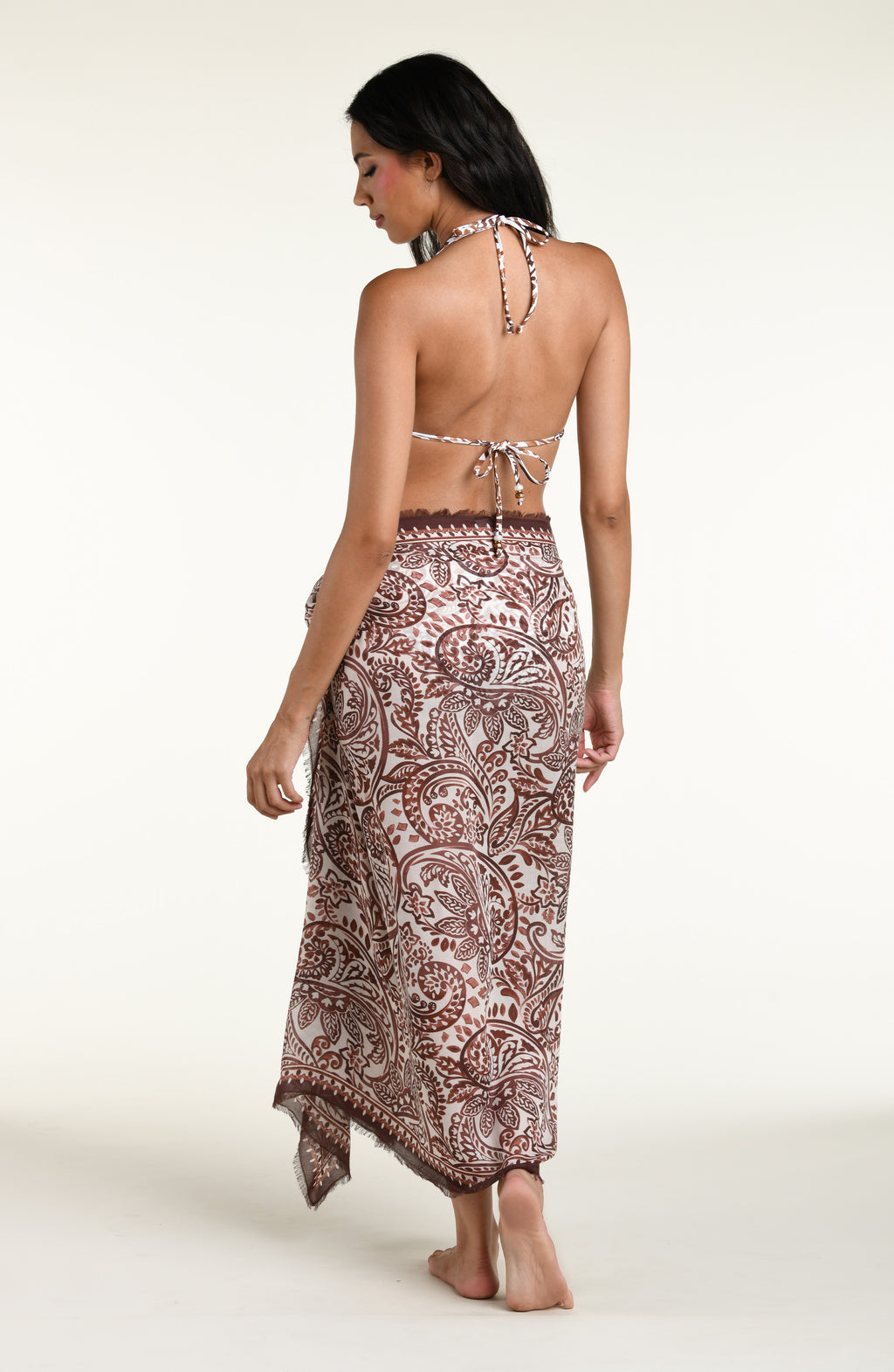 A woman modeling a halter bikini top and a matching high-slit sarong wrap skirt, both with a brown and beige paisley pattern on a white background, set against a plain light beige studio backdrop.