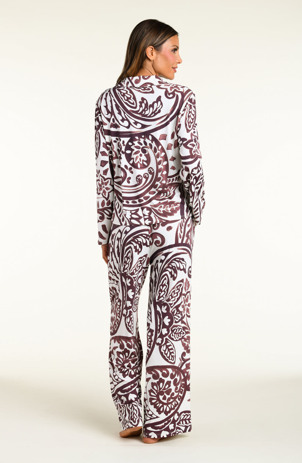 A woman modeling a matching long-sleeve button-front pajama top and wide-leg pajama pants set with an intricate paisley pattern in dark brown and white, set against a plain off-white studio background.