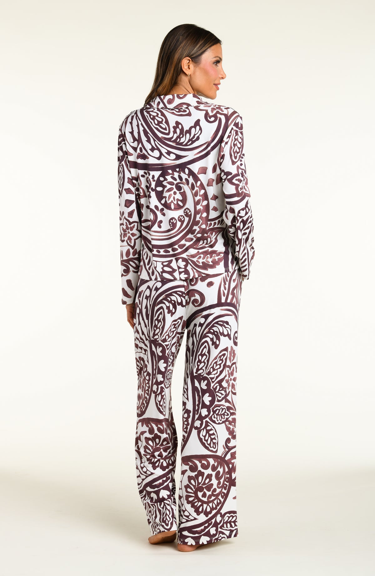 A woman modeling a matching long-sleeve button-front pajama top and wide-leg pajama pants set with an intricate paisley pattern in dark brown and white, set against a plain off-white studio background.