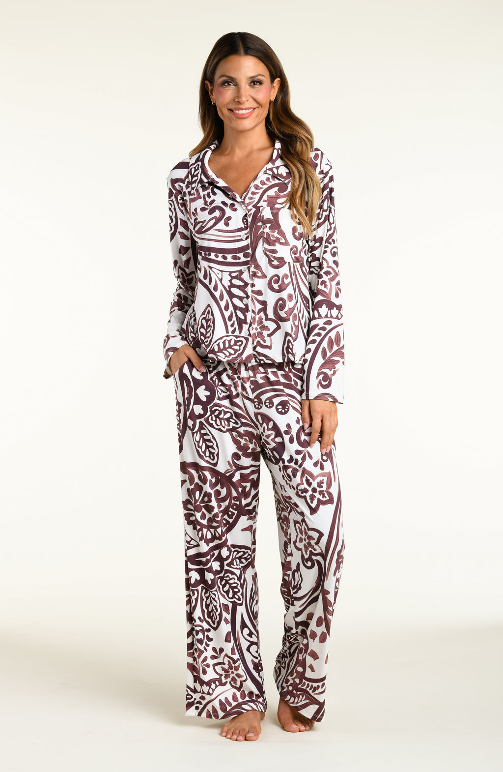 A woman modeling a matching long-sleeve button-front pajama top and wide-leg pajama pants set with an intricate paisley pattern in dark brown and white, set against a plain off-white studio background.
