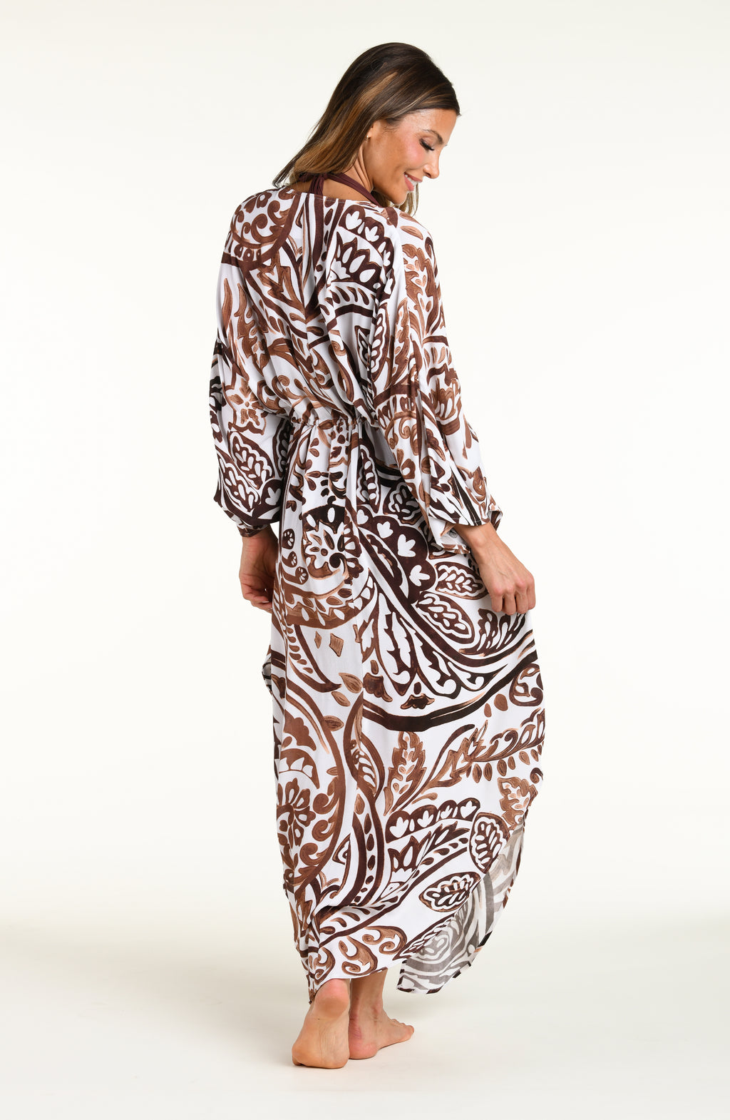 A woman modeling a long white caftan cover-up with a brown and white bohemian paisley pattern, standing against a plain white studio background.