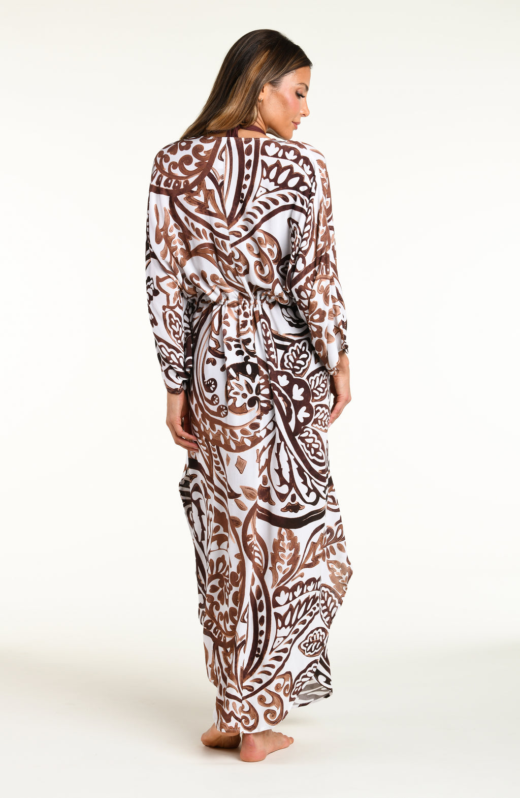 A woman modeling a long white caftan cover-up with a brown and white bohemian paisley pattern, standing against a plain white studio background.