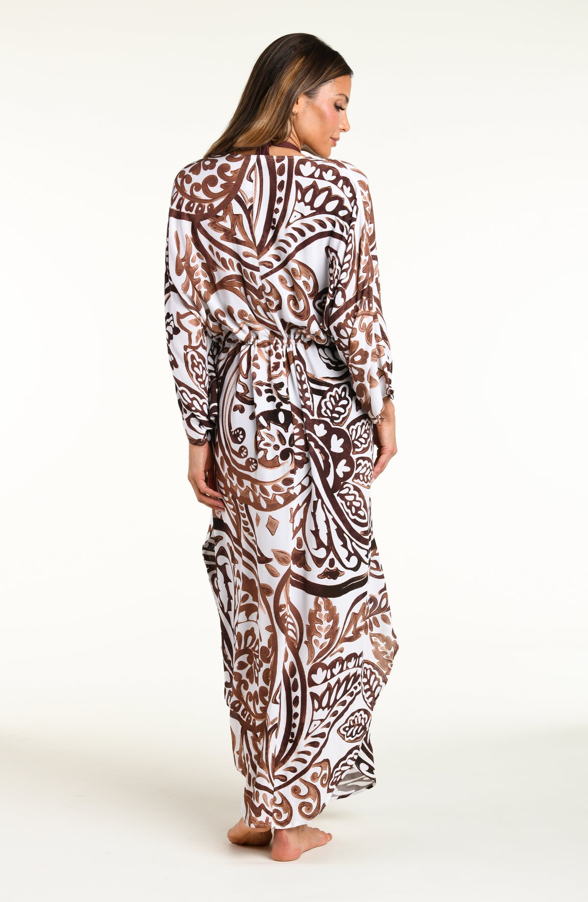 A woman modeling a long white caftan cover-up with a brown and white bohemian paisley pattern, standing against a plain white studio background.