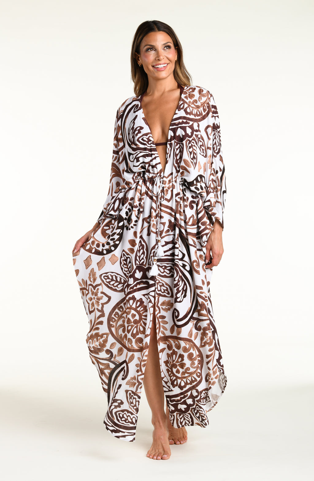 A woman modeling a long white caftan cover-up with a brown and white bohemian paisley pattern, standing against a plain white studio background.