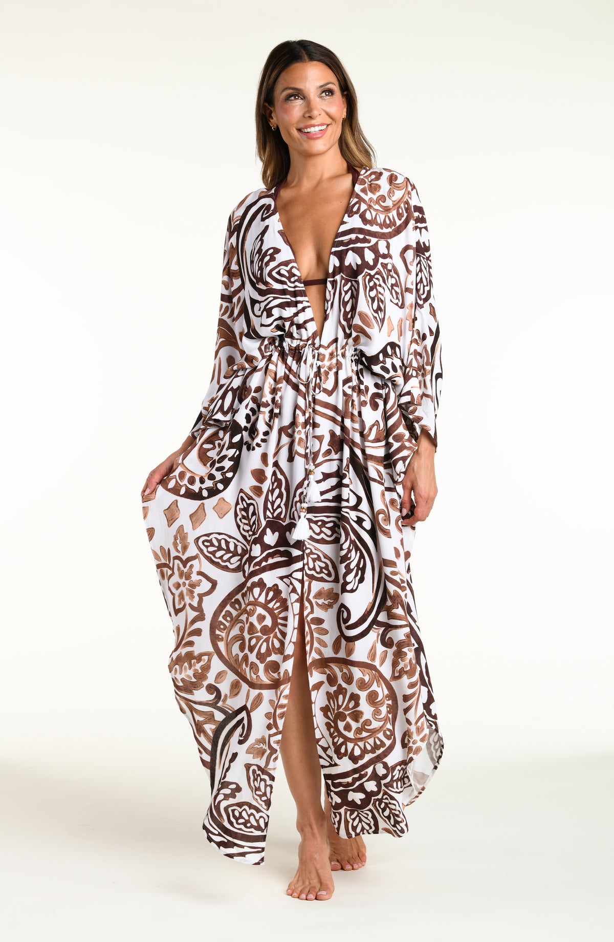 A woman modeling a long white caftan cover-up with a brown and white bohemian paisley pattern, standing against a plain white studio background.