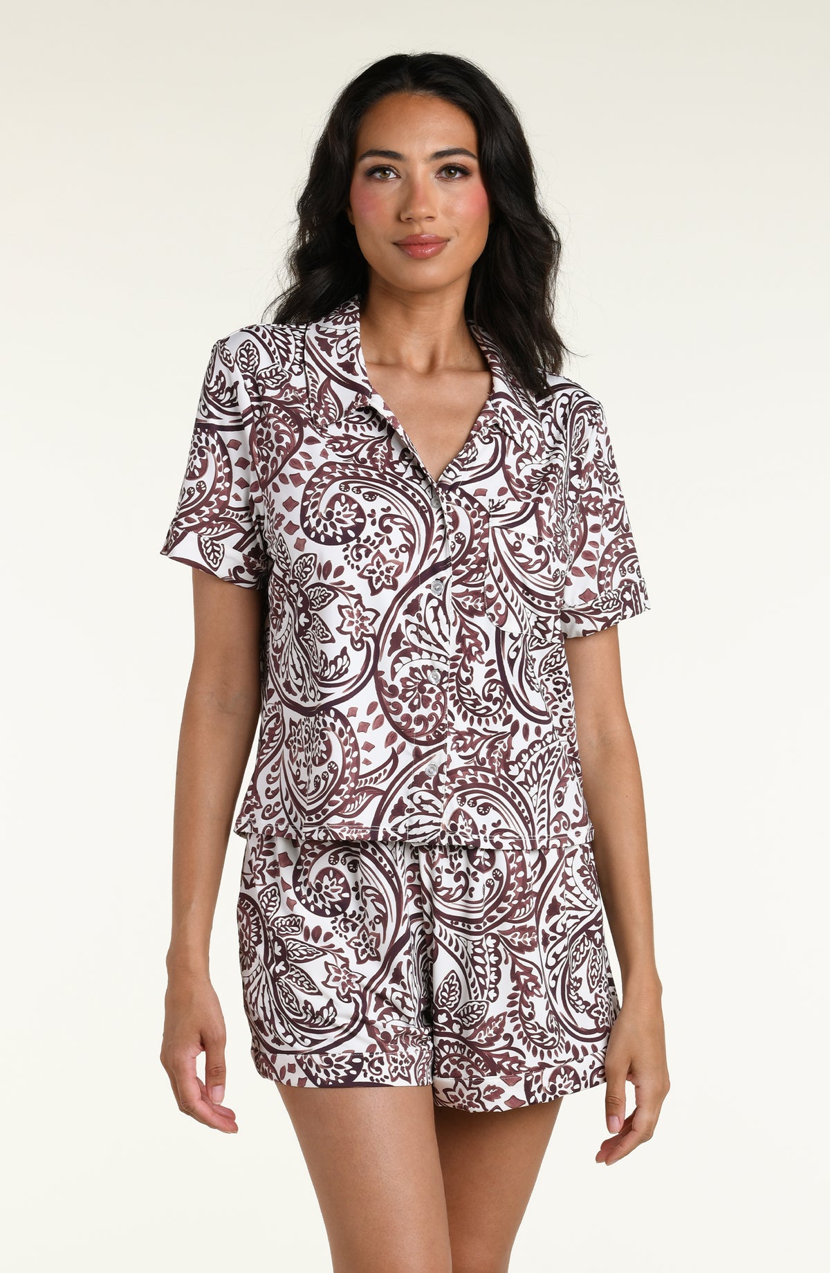 A woman modeling a pajama short-sleeve collared top and shorts set with an all-over brown paisley pattern on a white background, photographed against a plain light studio backdrop.