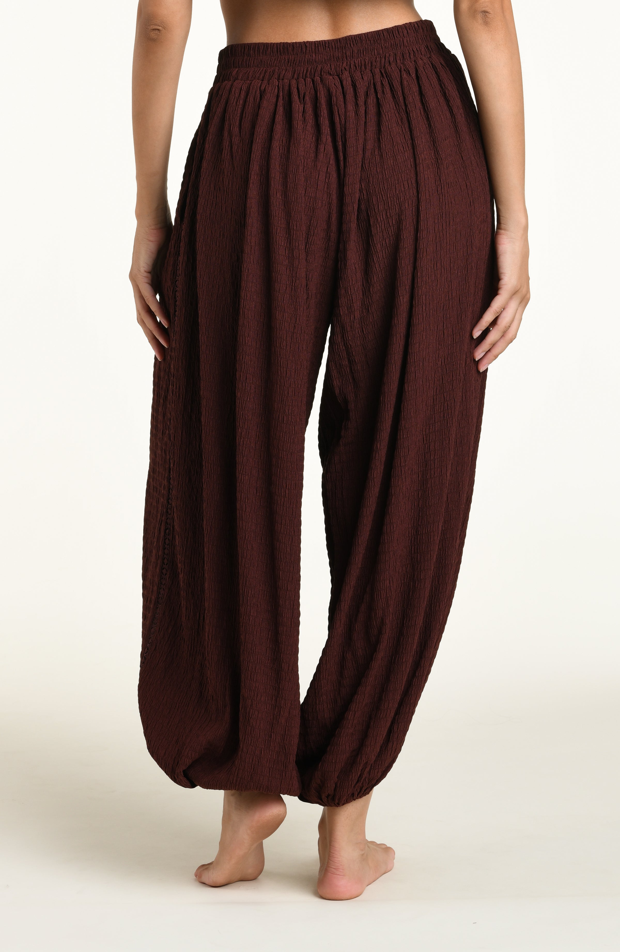 A woman wearing java brown colored crochet style pants against a white background. 