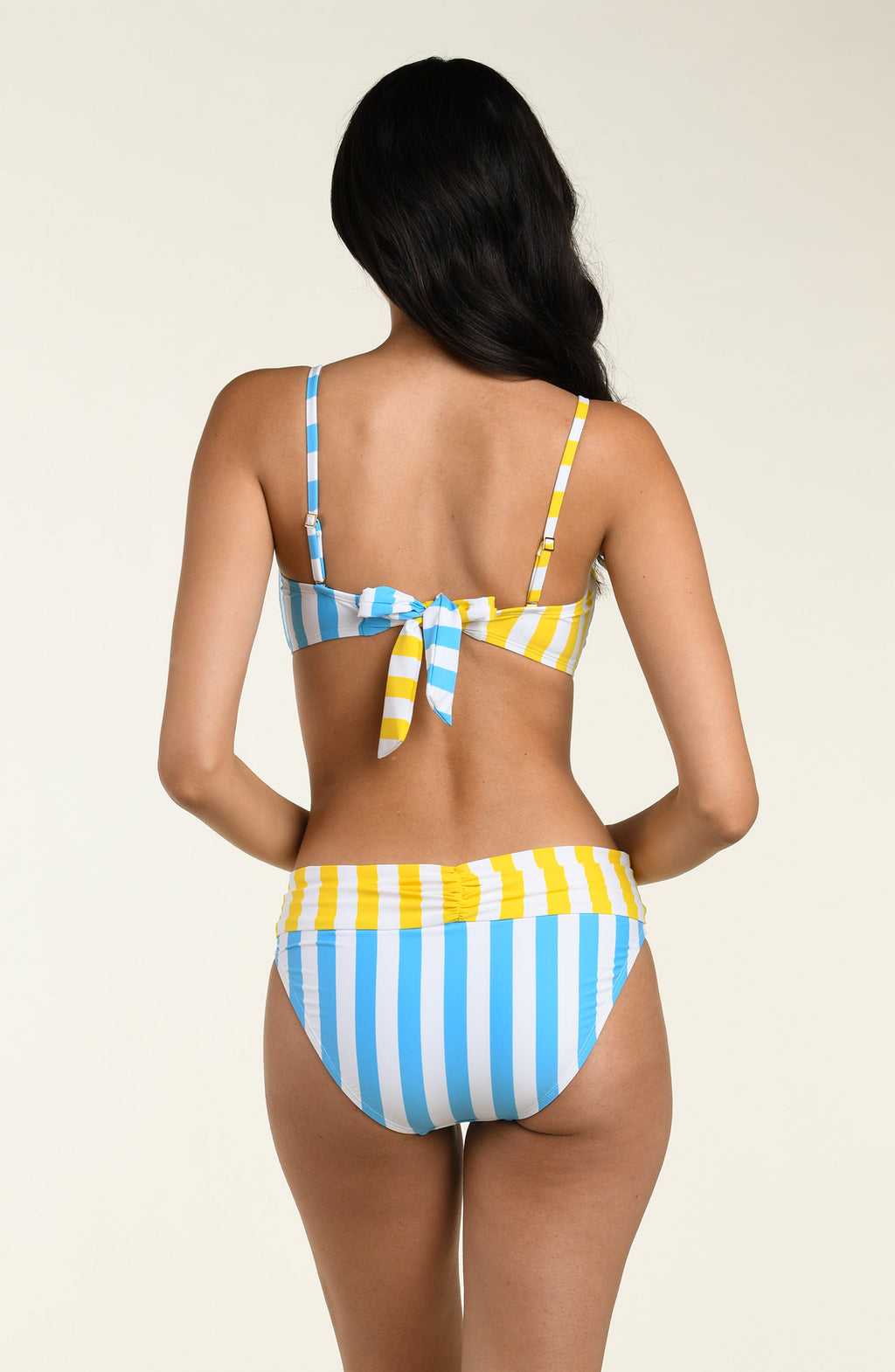 Woman wearing a yellow and blue striped bikini top and matching bottom on a beige background