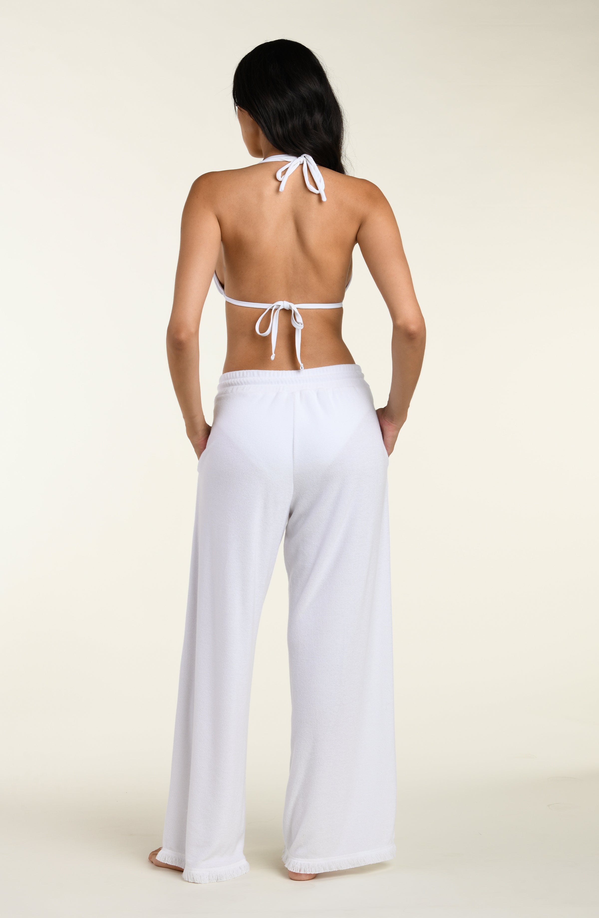 Woman wearing white cover up beach pants with a white bikini top against a white background. 