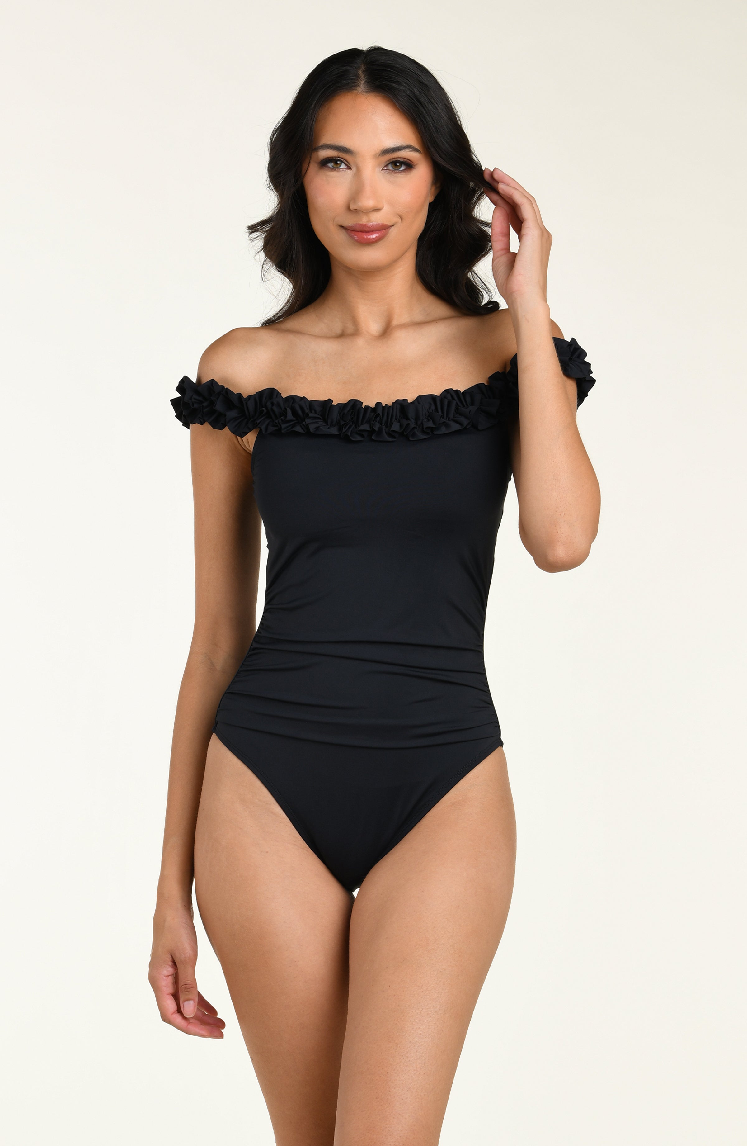 Woman wearing a black off-shoulder one piece swimsuit with ruffled details on a beige background