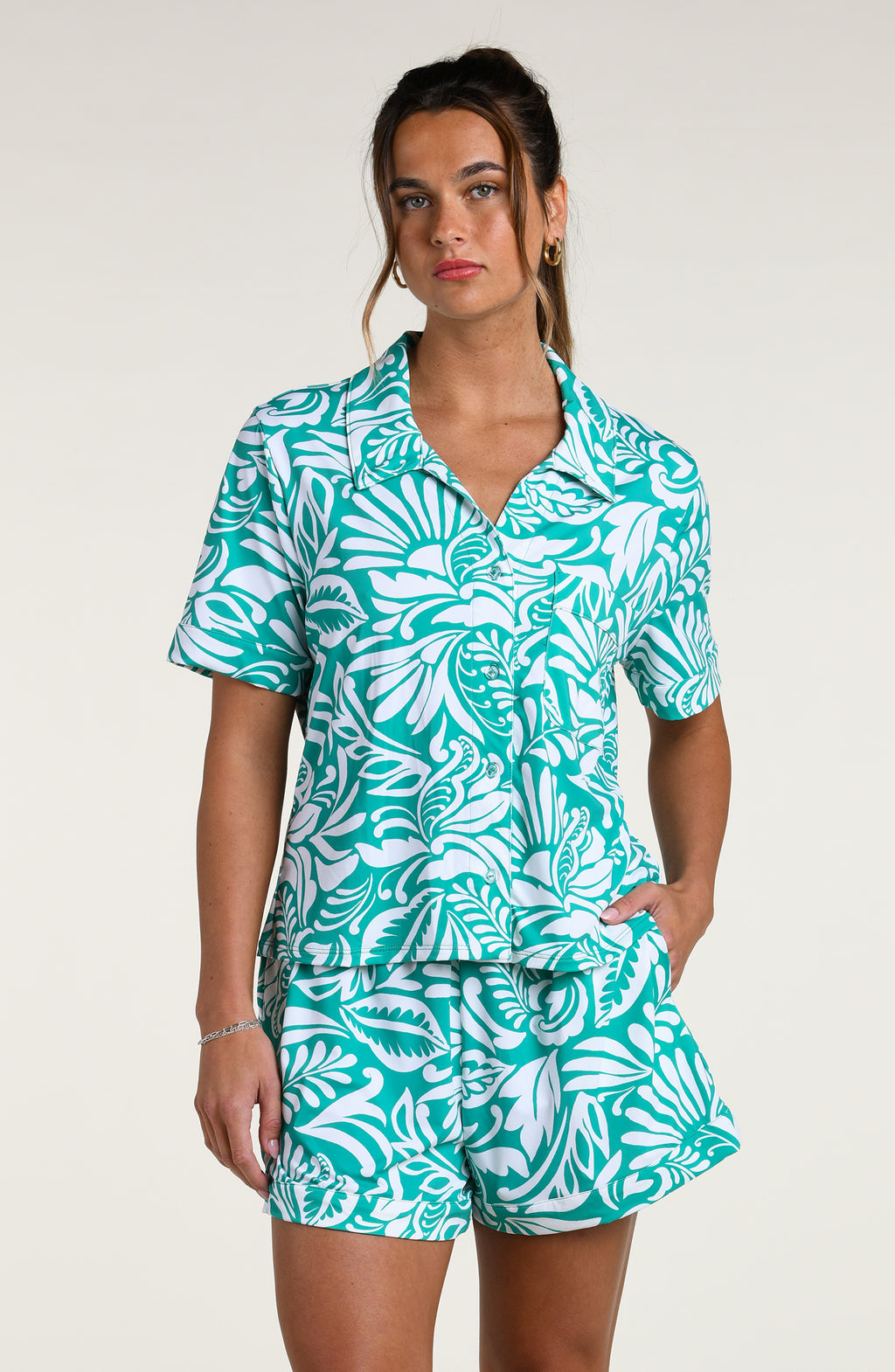 Woman wearing a teal and white patterned short sleeve shirt and shorts pajama set against a plain background