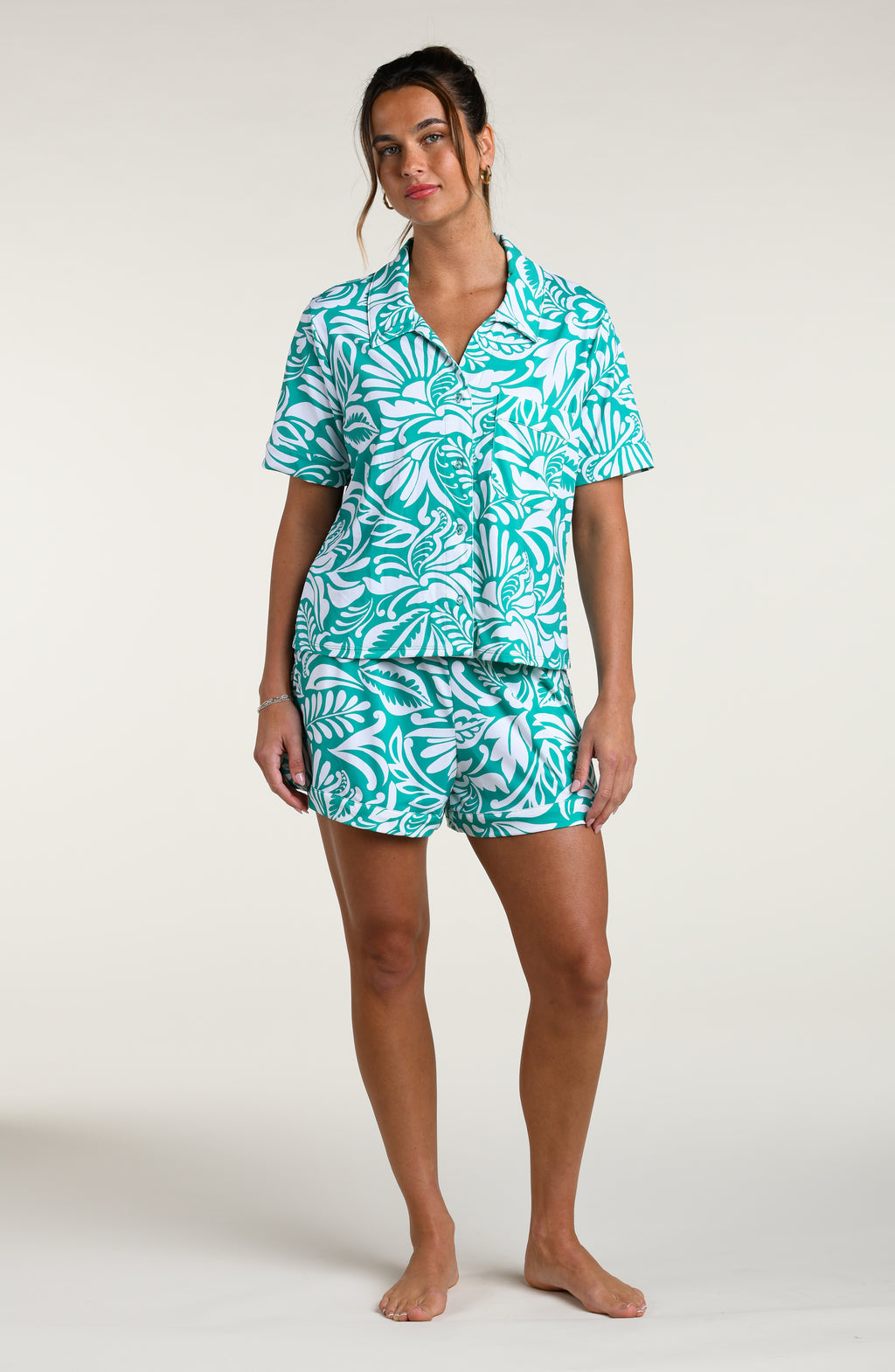 Woman wearing a teal and white patterned short sleeve shirt and shorts pajama set against a plain background