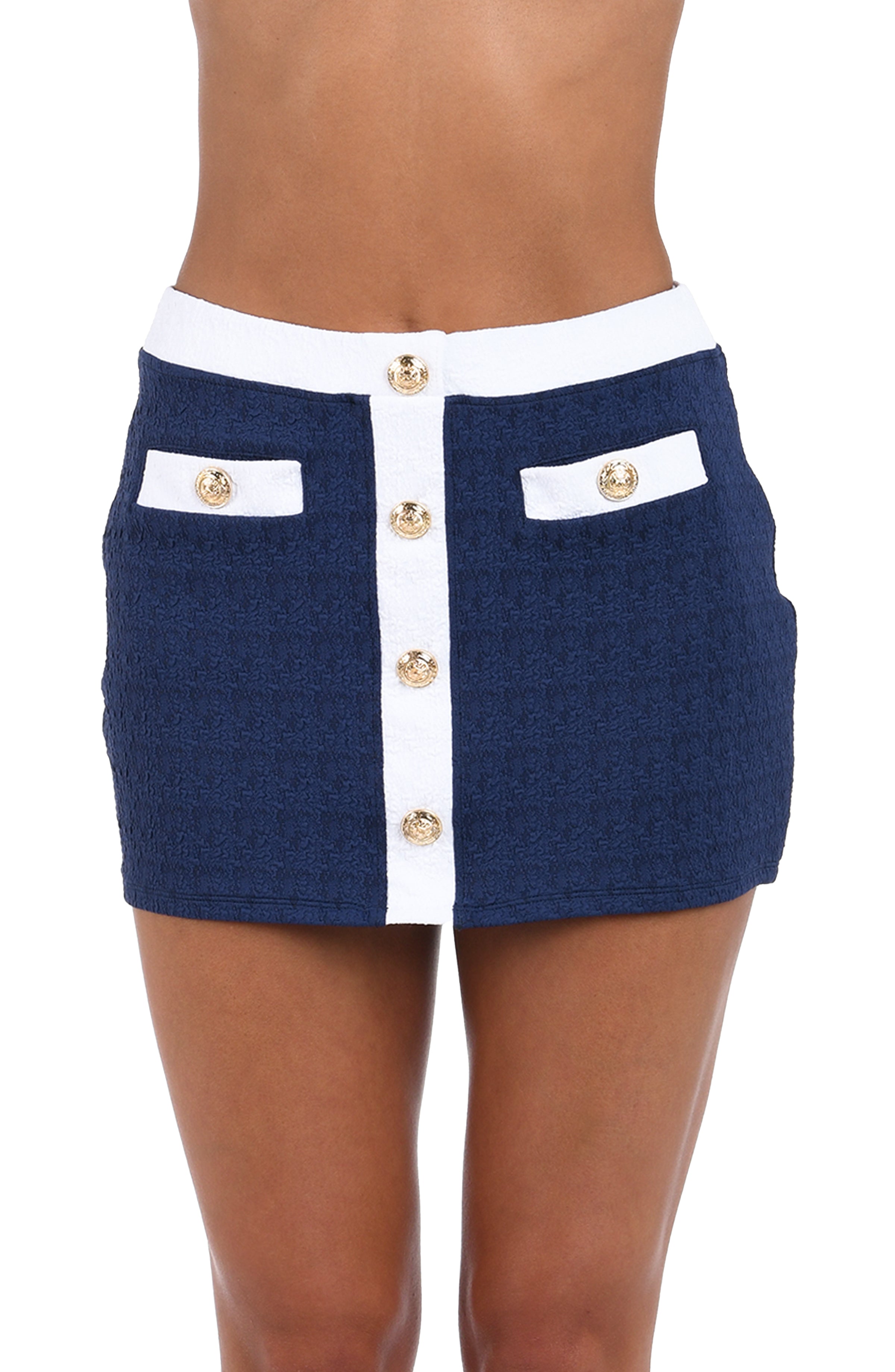 A close-up of a model wearing a navy blue textured tweed mini skirt with a white center panel, white waistband, and white pocket flaps accented with gold-tone buttons.