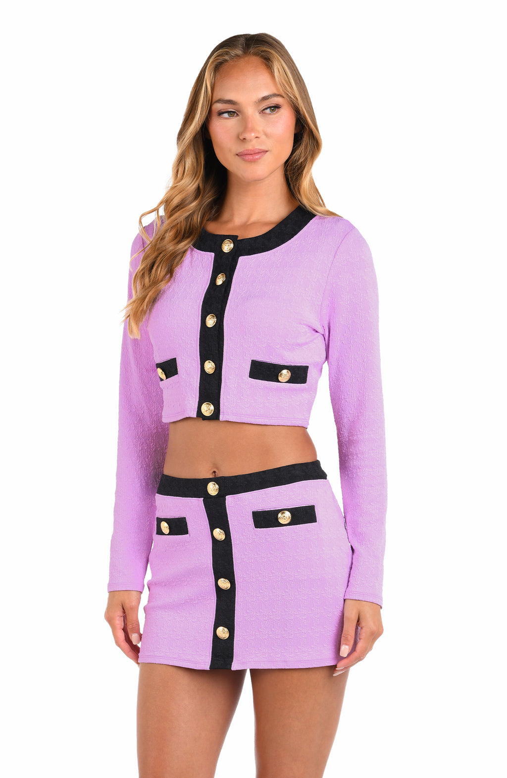A woman modeling a lavender knit two-piece set consisting of a cropped long-sleeve cardigan and a matching mini skirt, both featuring black trim and gold-tone decorative buttons.