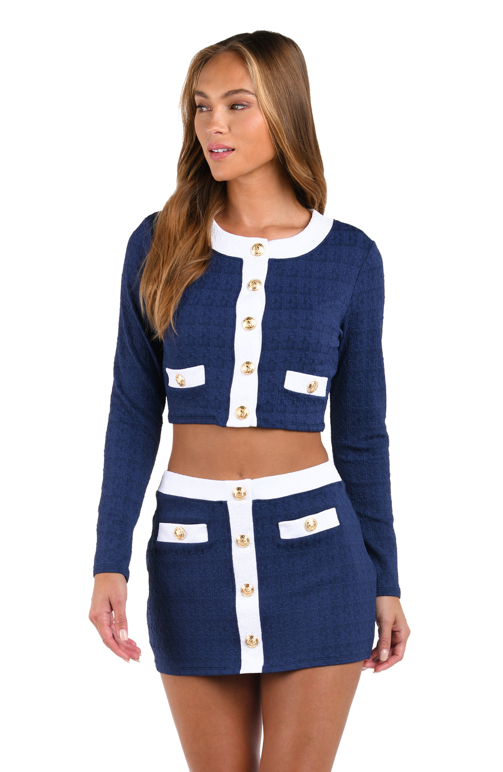A woman modeling a navy blue tweed two-piece set consisting of a cropped long-sleeve cardigan and a matching mini skirt, both featuring white contrast panels and gold-tone decorative buttons.