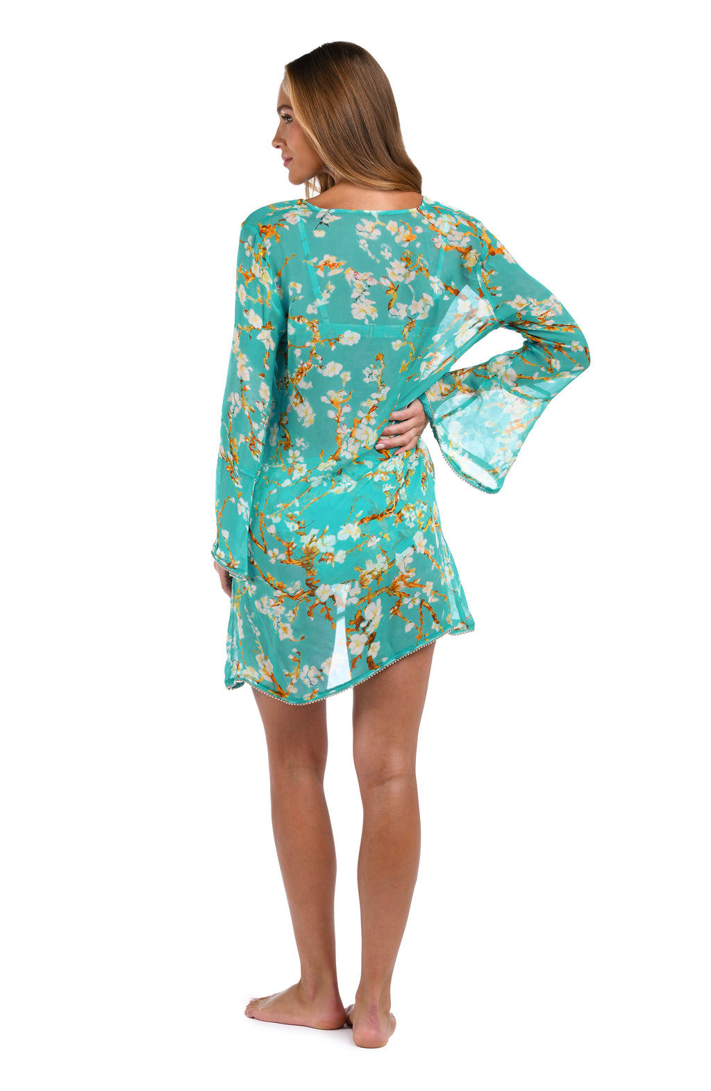 A woman modeling a turquoise blue long-sleeve tunic dress with a white and yellow floral print, shown from the back against a white background.