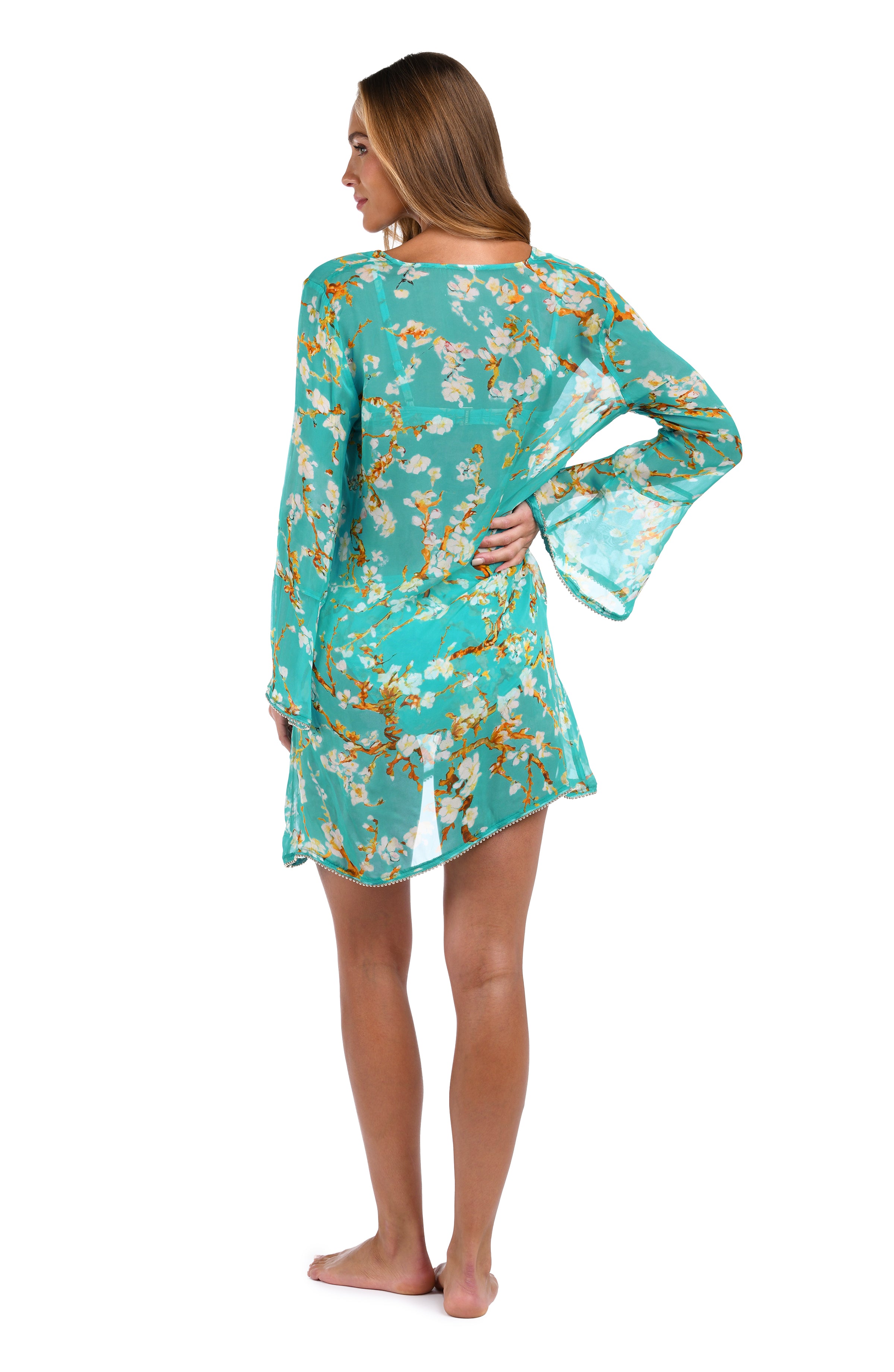 A woman modeling a turquoise blue long-sleeve tunic dress with a white and yellow floral print, shown from the back against a white background.