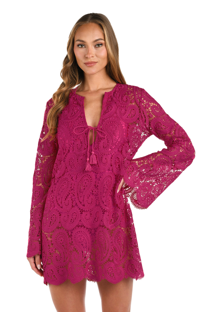 A woman wearing a fuchsia lace long-sleeve mini dress with a tie-neck detail, set against a plain white background.