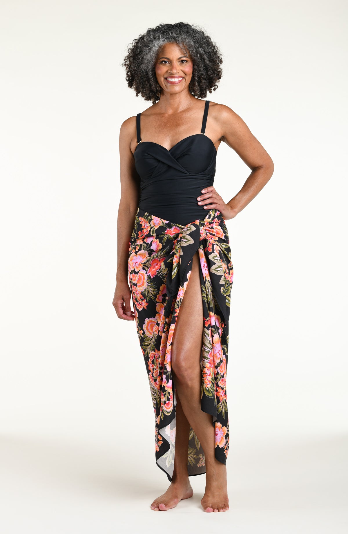 A woman modeling a black one piece swimsuit and a wrap sarong with a vibrant pink, orange, and green floral print, featuring a high side slit and a tied waist.