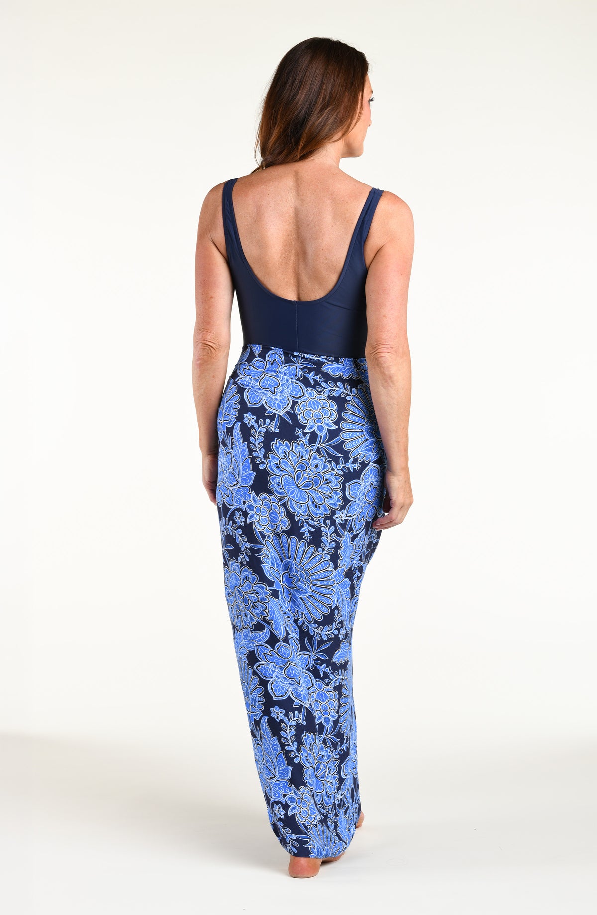 Woman wearing an indigo blue one piece swimsuit under an indigo blue multicolored sarong against a white background. 