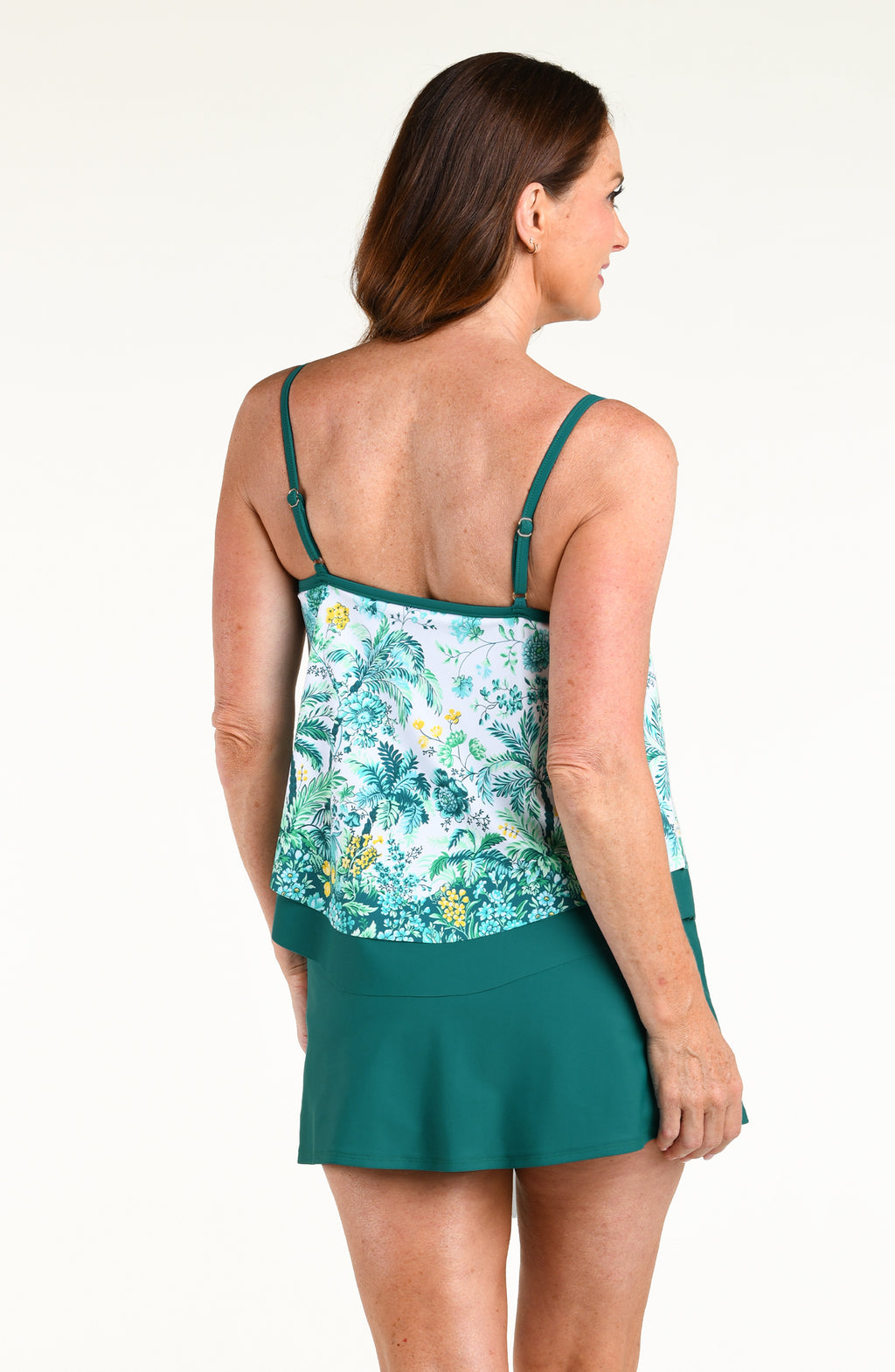 A woman modeling a tankini top featuring a tropical floral print and a solid teal swim skirt, set against a plain white studio background.