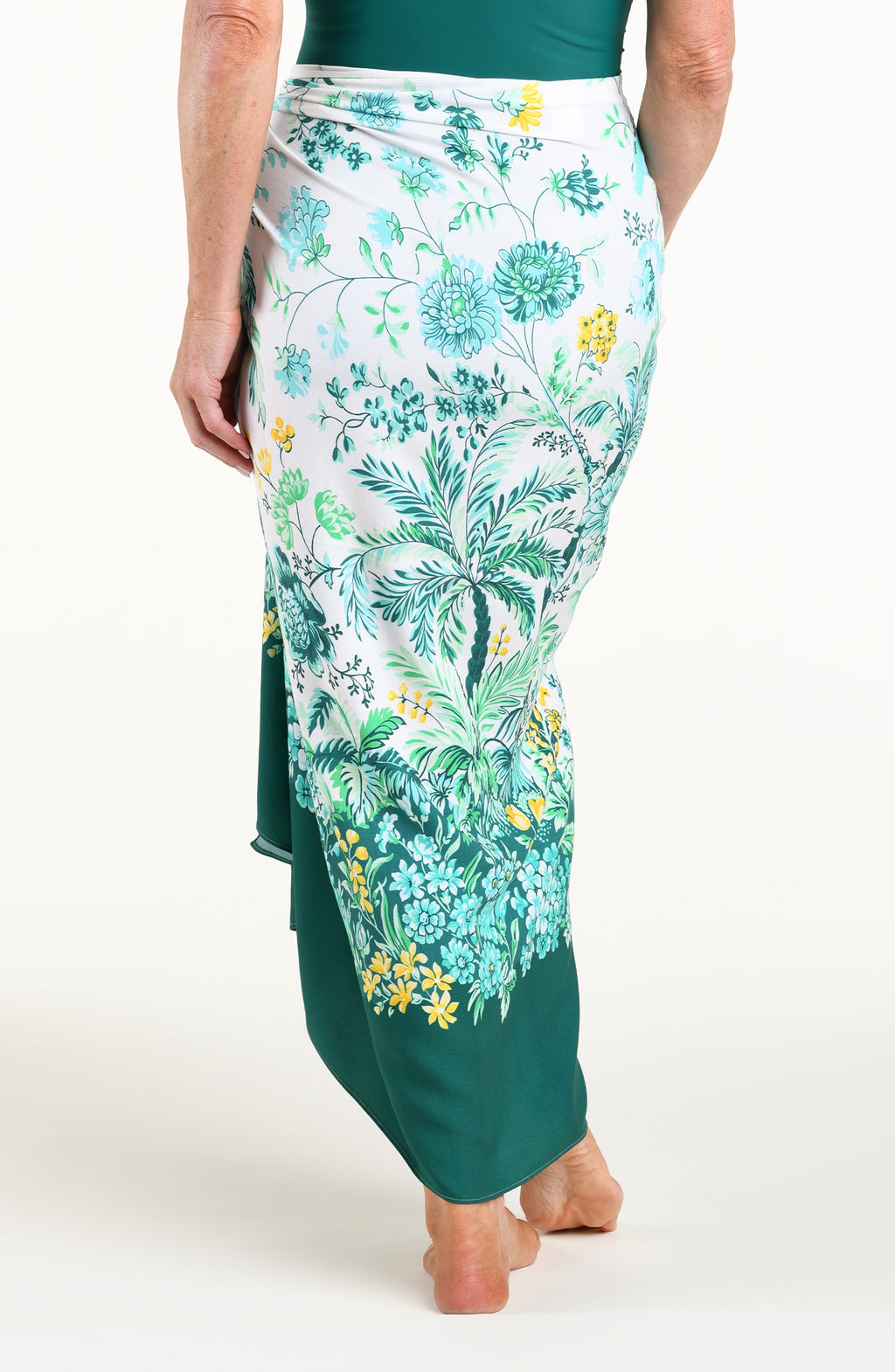 A woman wearing a tropical floral print sarong wrap skirt in green, blue, and yellow on a white base, shown from the back against a plain white studio background.