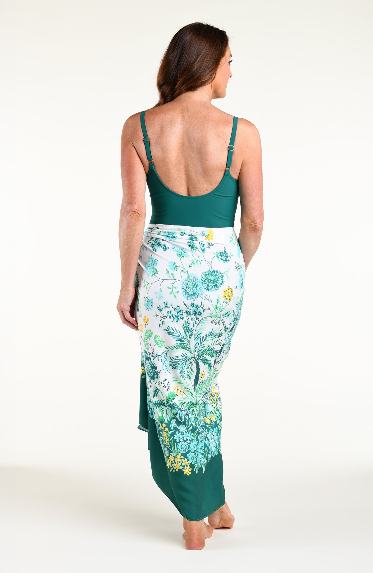 A woman modeling a solid teal green one piece swimsuit and a matching teal green tropical floral print sarong skirt, set against a plain white studio background.