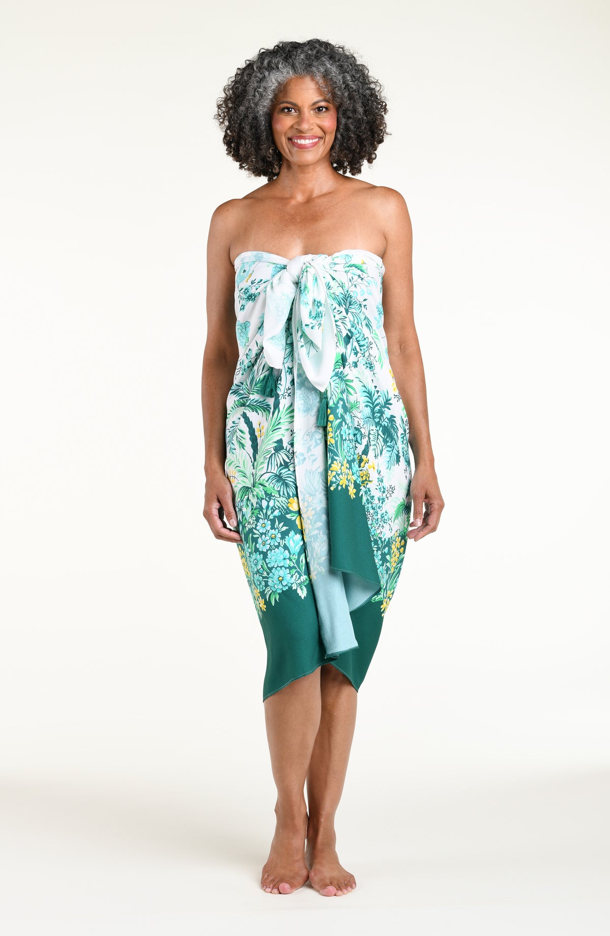 A woman models a high-low wrap sarong with a green and white botanical print, featuring tassel ties and a side slit, shown against a plain light background.
