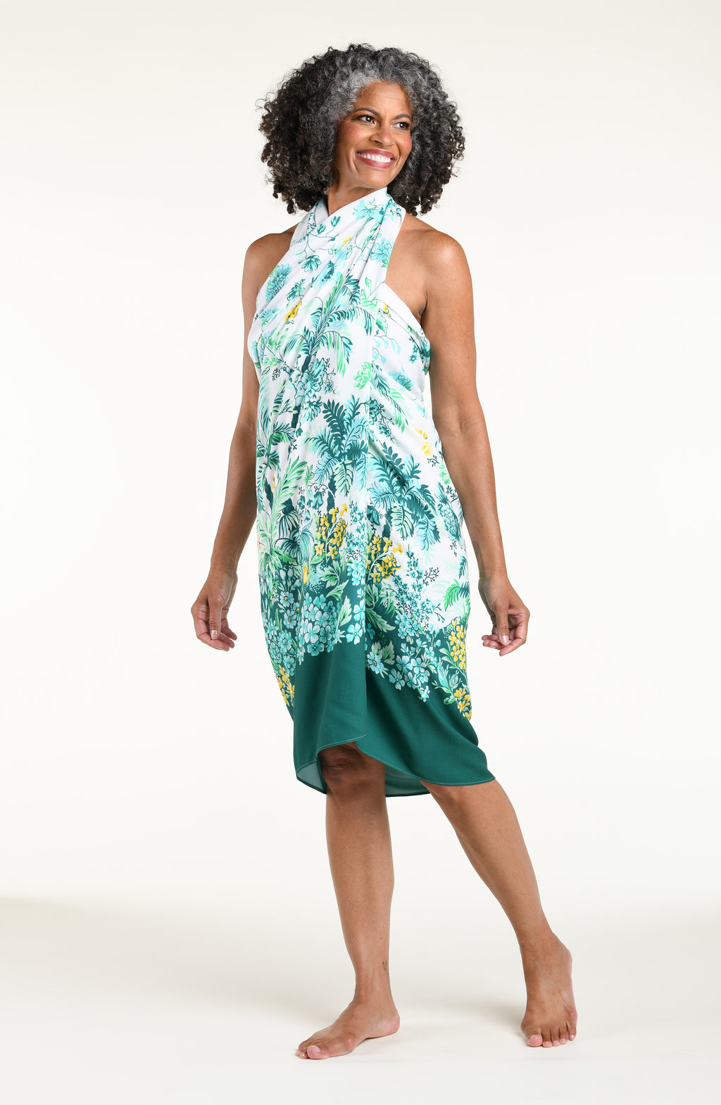 A woman models a high-low wrap sarong with a green and white botanical print, featuring tassel ties and a side slit, shown against a plain light background.