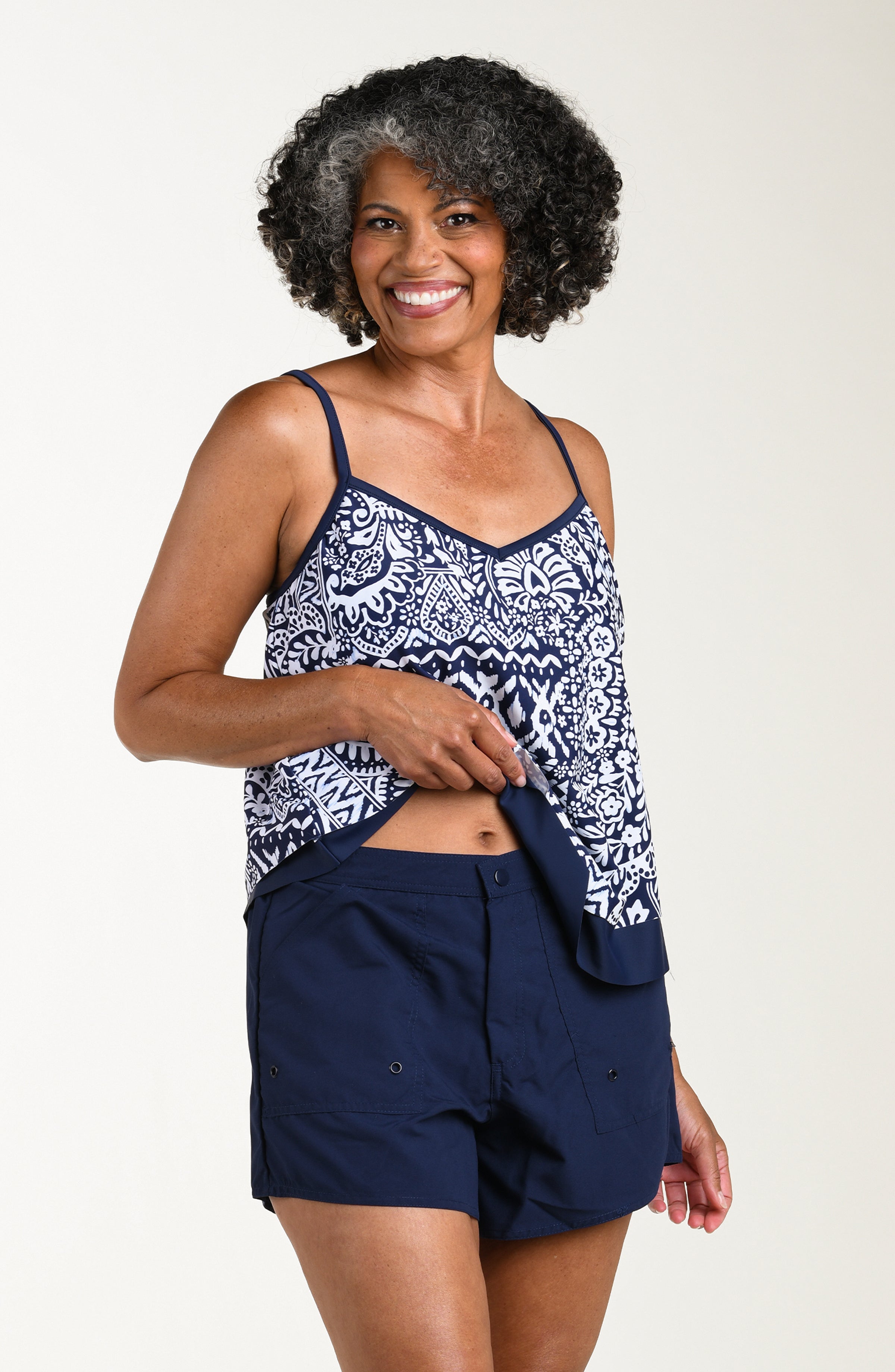 Front image of model wearing a multicolored navy blue paisley printed tankini top featuring thin adjustable straps, a V-neckline, and a flowy handkerchief hem. This is paired with matching colored board shorts.