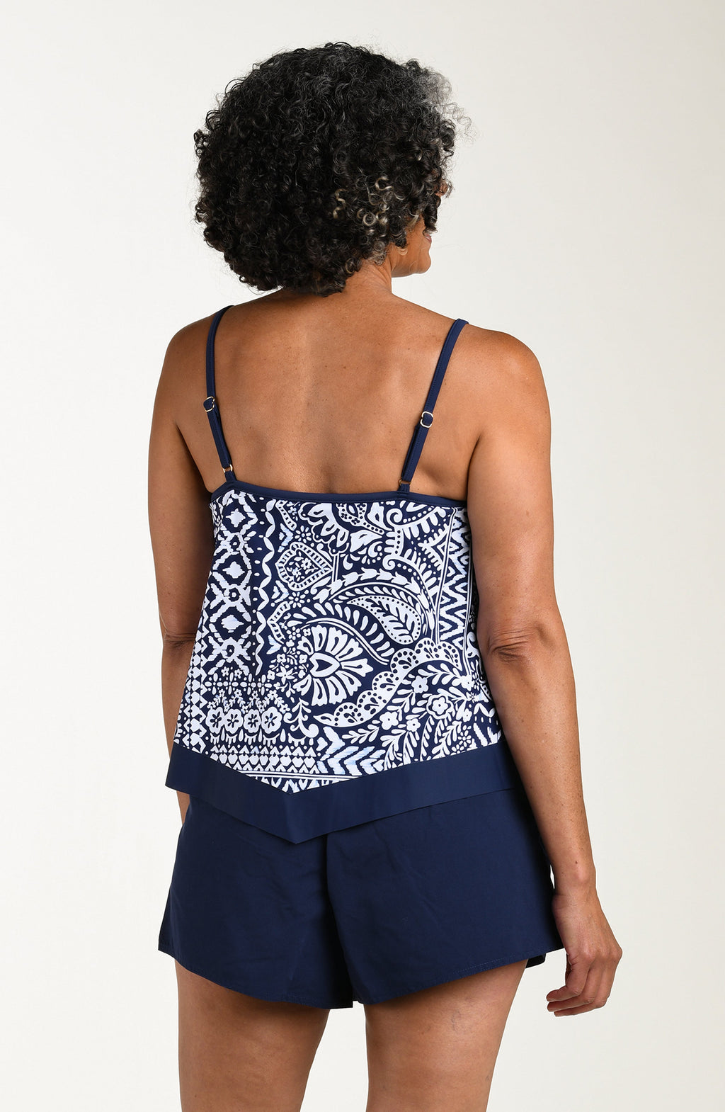 Back image of model wearing a multicolored navy blue paisley printed tankini top featuring thin adjustable straps, and a flowy handkerchief hem. This is paired with matching colored board shorts.