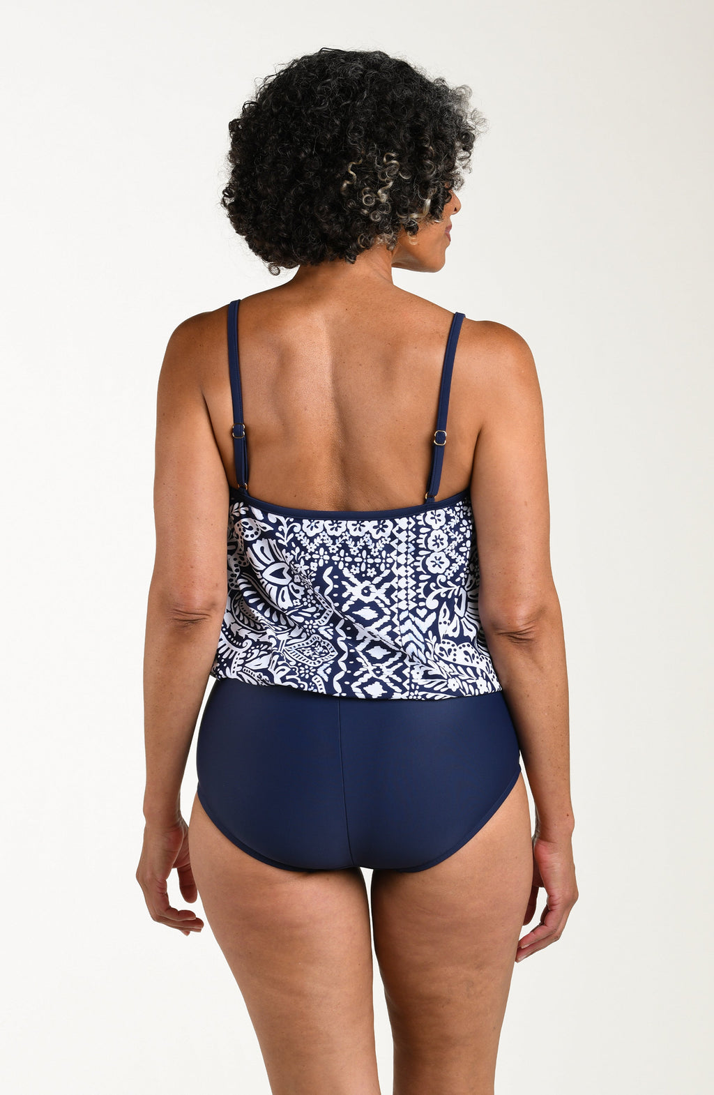 Back image of model wearing a multicolored navy blue paisley printed one-piece swimsuit featuring thin straps, a straight back, and a loose-flowing overlay. The top looks like a tankini and the bottom is solid indigo (navy blue) colored.