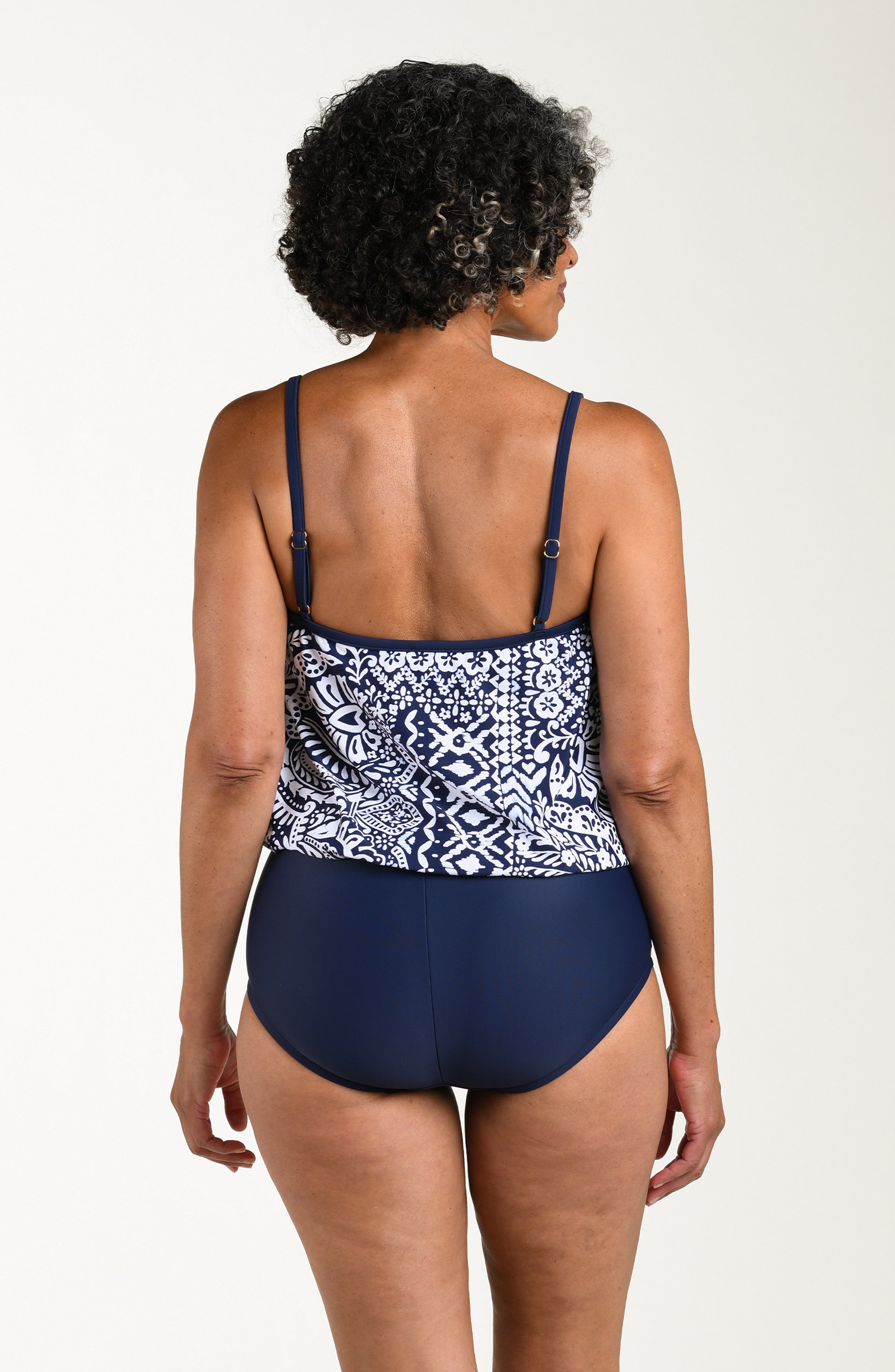 Back image of model wearing a multicolored navy blue paisley printed one-piece swimsuit featuring thin straps, a straight back, and a loose-flowing overlay. The top looks like a tankini and the bottom is solid indigo (navy blue) colored.