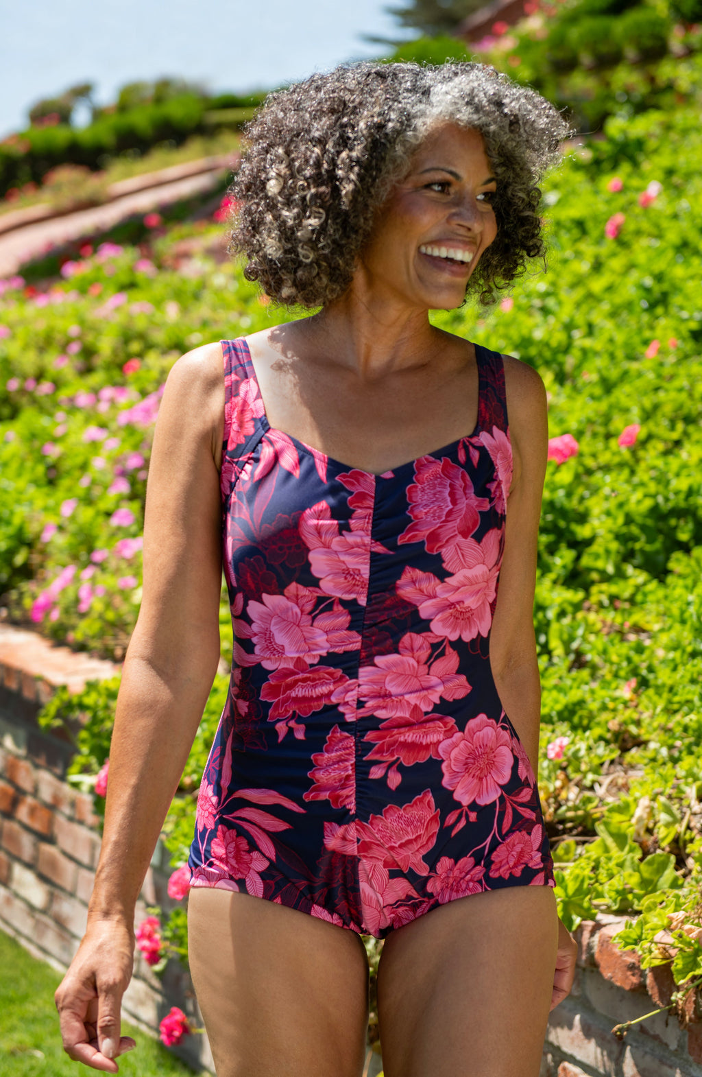 Woman wearing a floral swimsuit standing in a garden with pink flowers and greenery.
