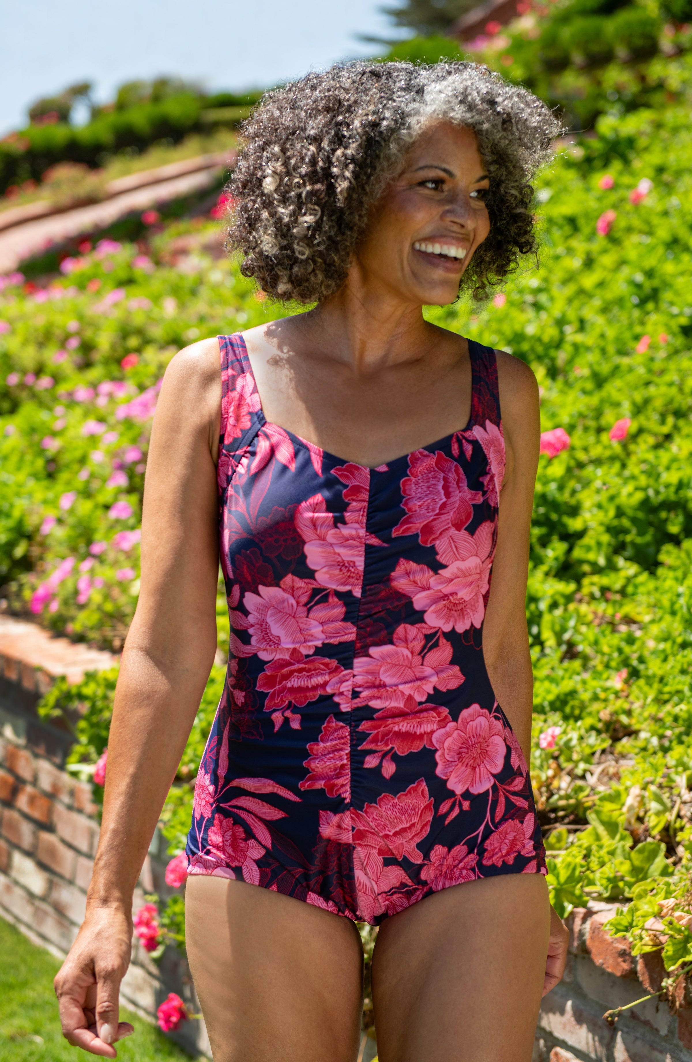 Woman wearing a floral swimsuit standing in a garden with pink flowers and greenery.
