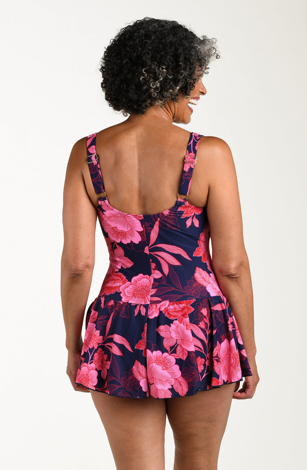Back image of model wearing a multicolored (pink) floral printed one-piece swim dress with wide straps, a scoop neckline, flared skirt, and full rear coverage.