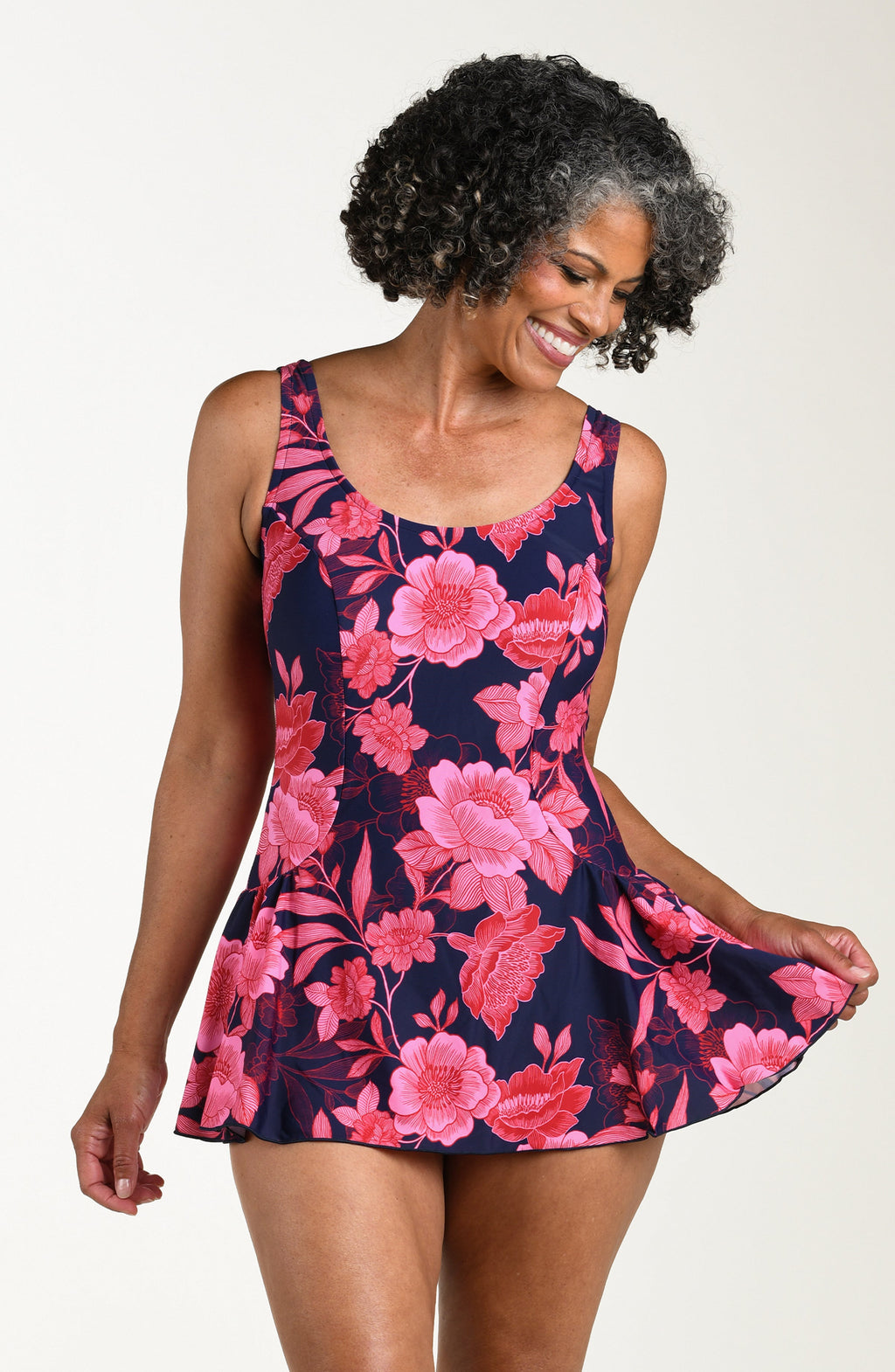 Front image of model wearing a multicolored (pink) floral printed one-piece swim dress with wide straps, a scoop neckline, and a flared skirt.