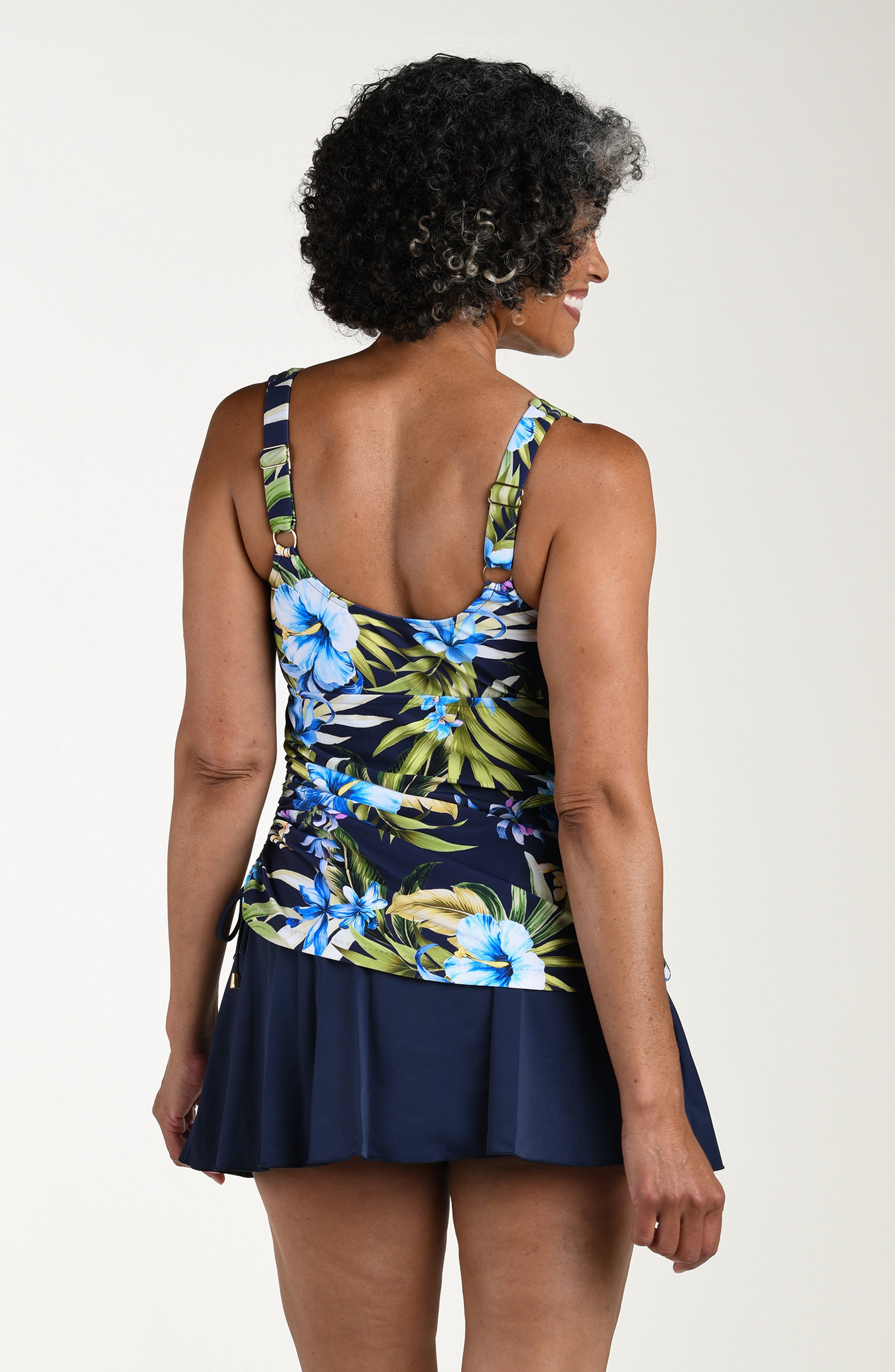 Back image of model wearing a multicolored (blue) tropical swim dress featuring adjustable over the shoulder straps and side tie detail. The top looks like a tankini and the bottom is solid indigo skirt.